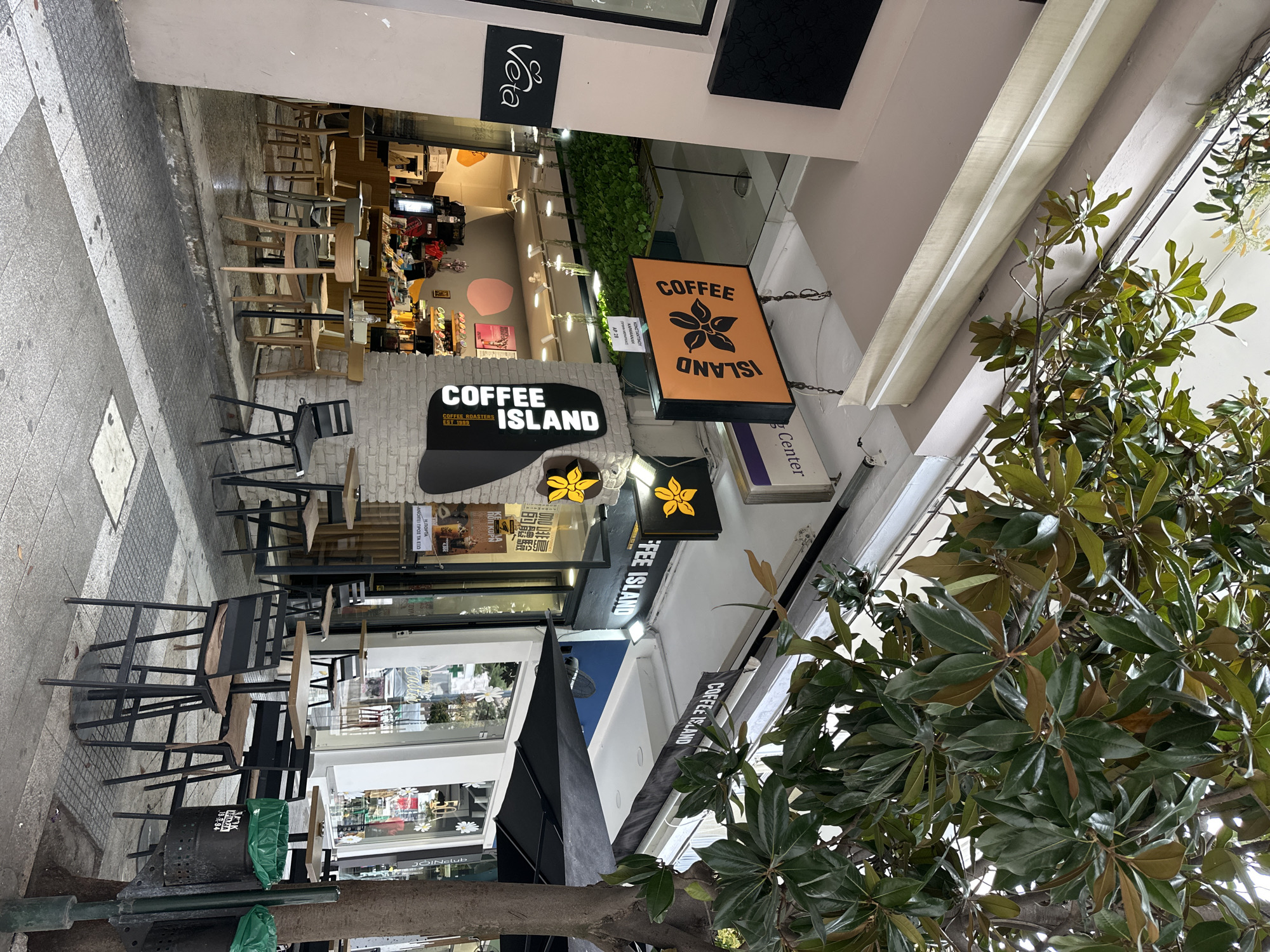 The image is a street scene featuring a Coffee Island store in Serres, Greece. Several signs for Coffee Island are visible, one with a flower logo and another that says "Coffee Roasters EST 1999". Adjacent to the store are various retail establishments, including one advertising "ΚΩΝΣΤΑΝΤΙΝΟΥ ΚΑΡΑΜΑΝΛΗ ΑΡ 28". Outdoor seating is arranged on the sidewalk, and nearby there's a trash can with a green bag and a leafy tree.