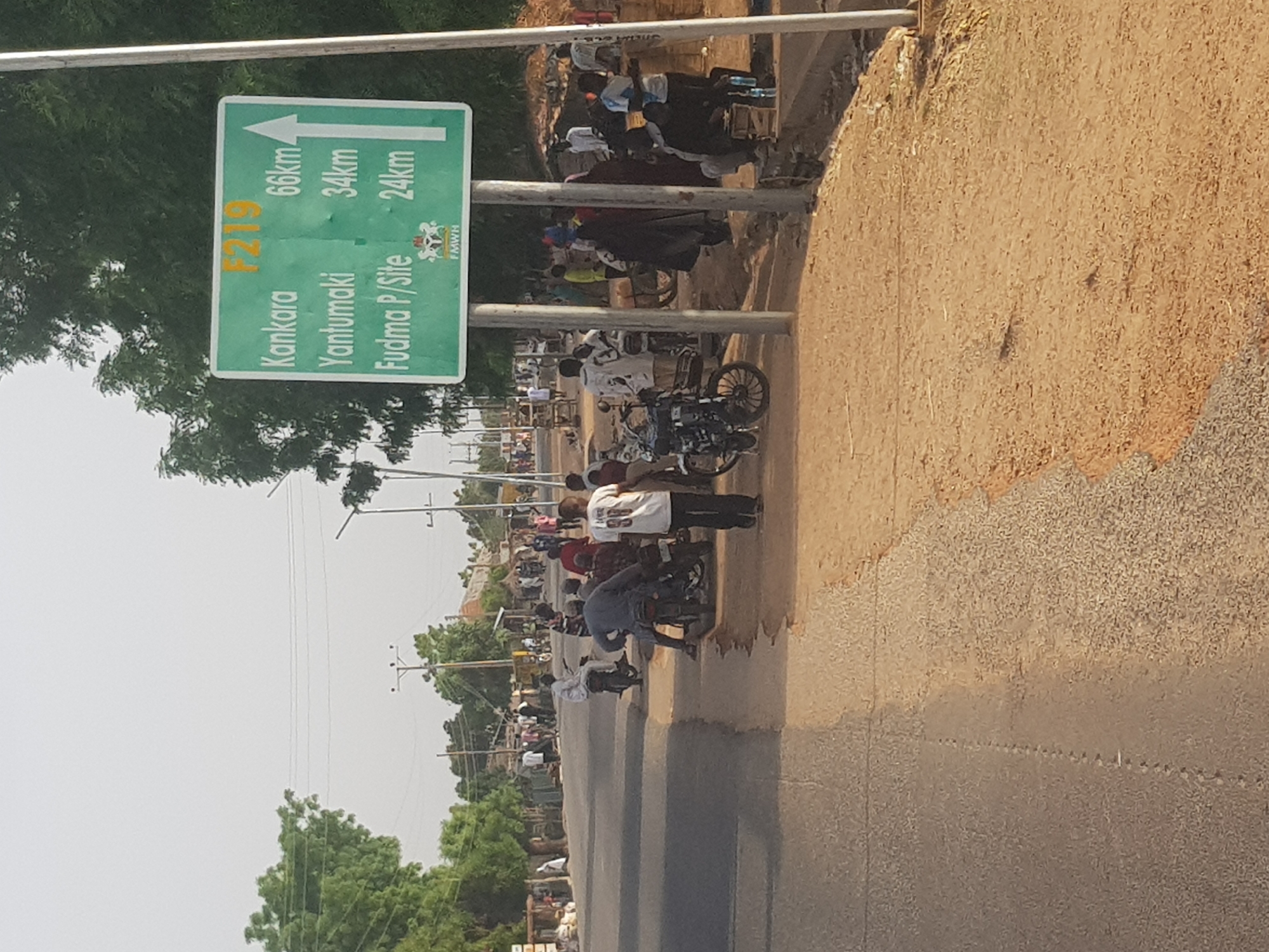 The image shows a roadside scene in Dabawa, Nigeria. A green sign indicates distances to Kankara (66km), Yantumaki (34km), and Fudma P/Site (24km). People and motorcycles are gathered alongside the road. The road is paved, with dirt and gravel on the shoulder. Trees and power lines are visible in the background. The sky appears overcast.
