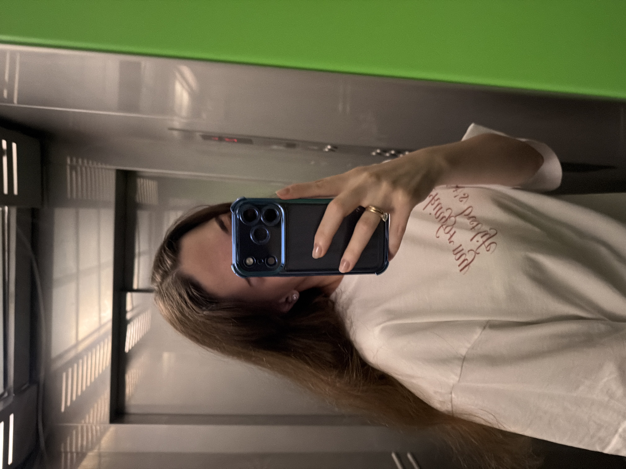 This is a selfie taken in what appears to be an elevator or a similar enclosed space with reflective surfaces. The primary subject is a young woman, seen in a reflection. She has long, brown hair and is wearing a white t-shirt with some red lettering on it. She is holding a dark-colored smartphone with a blue case in her right hand, using it to take the photo. A ring is visible on her right ring finger.
The background is a reflection of the elevator's interior, showing metallic walls and a green ceiling at the top of the frame. The lighting suggests it could be daytime, as there are reflections of what might be windows or light fixtures creating a striped pattern on the wall. The overall mood is casual and typical of a self-taken photograph. The visible text on her shirt is in a script font, but it is partially obscured and difficult to read clearly. The location is noted as Kyiv, Ukraine.
