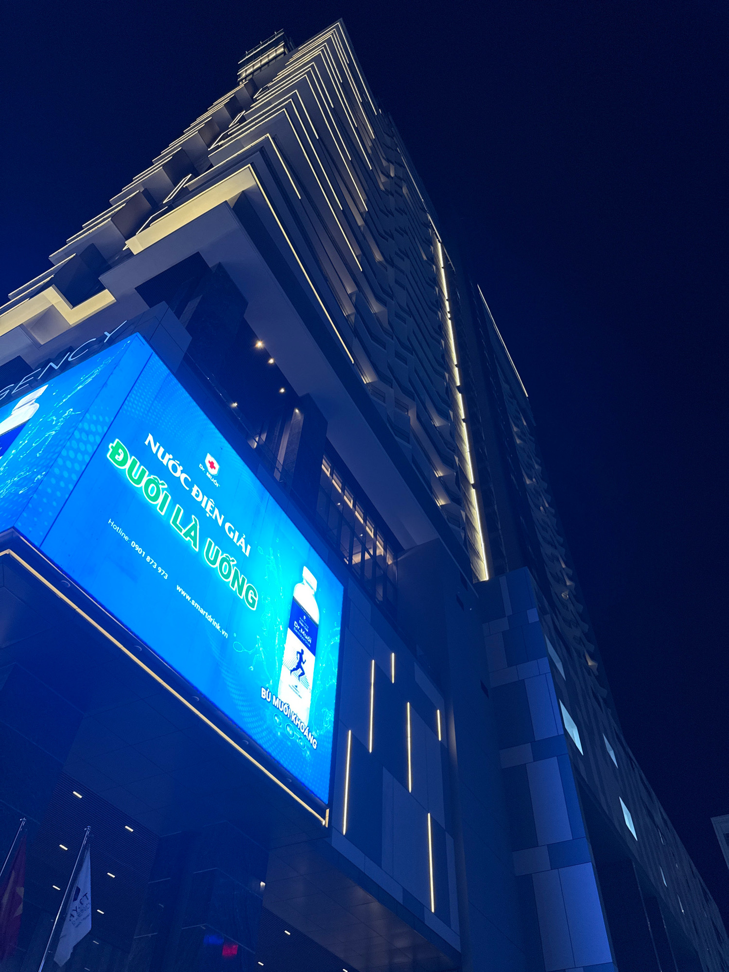 The photograph depicts a modern, illuminated high-rise building at night in Nha Trang, Vietnam. The building's facade features a striking architectural design with angular projections and horizontal lighting strips. A large, vibrant blue LED screen is prominently displayed on the lower section of the building, showcasing a product advertisement. The advertisement features Vietnamese text, a logo with a red cross, and an image of a bottle with a silhouette of a person. Below the main advertisement, a phone number and website address are visible. Two flags are positioned in the lower left corner of the image. The sky above is a deep, dark blue, indicating nighttime. The perspective is from a low angle, looking upwards towards the imposing structure.