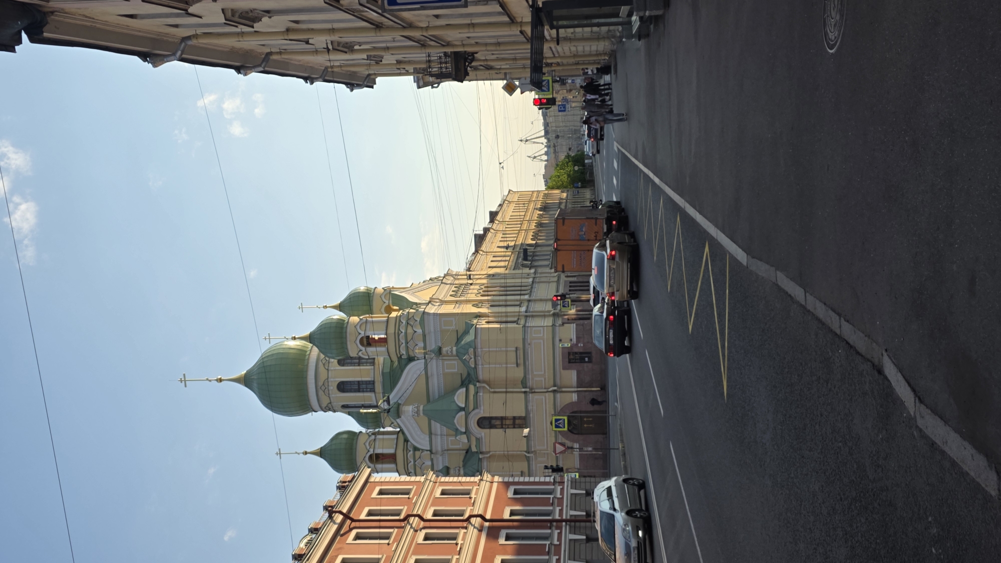 The image depicts a street scene in Saint Petersburg, Russia. On the left is a multi-story building with green domes and gold crosses, characteristic of Russian Orthodox churches. Adjacent to it is a building with red and orange brick facades. On the right is a multi-story building with a beige facade. The street is paved and has a lane divider. Cars are present on the street, and pedestrians can be seen near the far end of the street. The sky is blue with some clouds. Overhead lines are visible between the buildings. A traffic light is also visible at the end of the street.