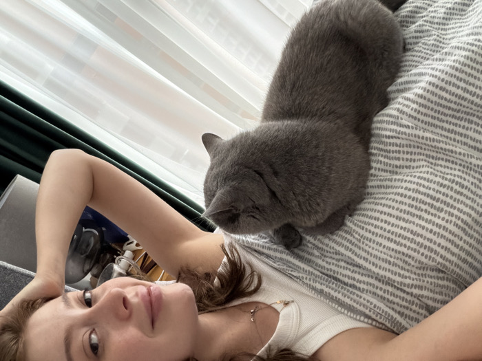 The image depicts an indoor scene in the city of Tbilisi, Georgia, featuring a young adult female and a solid grey cat, likely a British Shorthair. The female, positioned in the lower half of the frame, is lying on her back, looking directly at the camera. She has medium-length brown hair and fair skin, wearing a white ribbed tank top and a delicate gold-colored necklace. Her left arm is bent, with her hand resting near her head. The grey cat lies in the upper half of the frame, partially on the female's body or closely adjacent on a bed covered with a light-colored duvet featuring a grey dotted stripe pattern. The cat is oriented with its head towards the left, facing away from the camera. In the background, a large window with white sheer curtains and a dark green curtain panel is visible on the left, allowing natural light into the room. A dark grey headboard or armrest is partially visible on the far left, alongside indistinct household items on a surface.