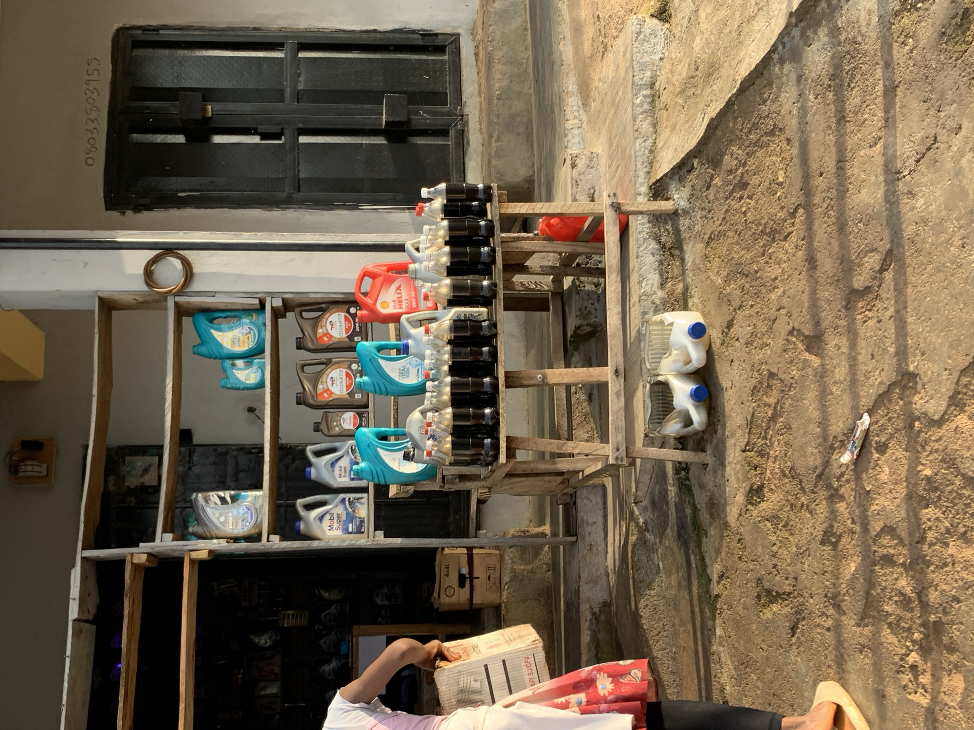 The image depicts an outdoor roadside stall in Jos, Nigeria, likely a small business selling automotive fluids and possibly other items. In the foreground, the ground is a mix of dirt and concrete, with some debris visible. To the left, a person is partially visible, wearing a pink and white top, a floral skirt, and beige slippers. They are holding a cardboard box.
The stall itself is constructed from wood and appears to have shelves stacked with various containers of automotive oil. Brands like "Mobil Super" and "QUARTZ" are discernible. In front of the shelves, a wooden table is set up, displaying rows of dark liquid in clear plastic bottles, capped with red and white. Behind the table, a red plastic container and a white plastic jug with a blue cap are visible on the ground. A metal door with a handle is set into a concrete building behind the stall, with a phone number, "08033803955," scrawled in dark ink above it. The overall impression is of a humble, functional commercial space operating in an urban or semi-urban environment.