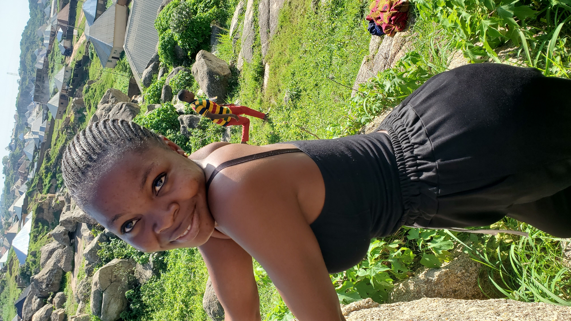 The media file captures a candid portrait of a young Black woman with braided hair, who is smiling at the camera. She is wearing a black tank top and black pants with a cinched waistband. The setting appears to be outdoors on a rocky, grassy hillside overlooking a village with houses and buildings scattered across a sloped landscape. In the background, another young person, possibly a child, is visible wearing a red and yellow striped shirt and red pants, adding a dynamic element to the scene. A piece of colorful fabric, possibly a wrapper, lies on the ground near some greenery. The lighting suggests it is daytime, and the overall mood is relaxed and cheerful, with the main subject exuding a warm smile. The location context of Jos, Nigeria, provides a geographical anchor for the scene, hinting at the type of landscape and architecture that might be expected in that region. No visible text is present in the image.