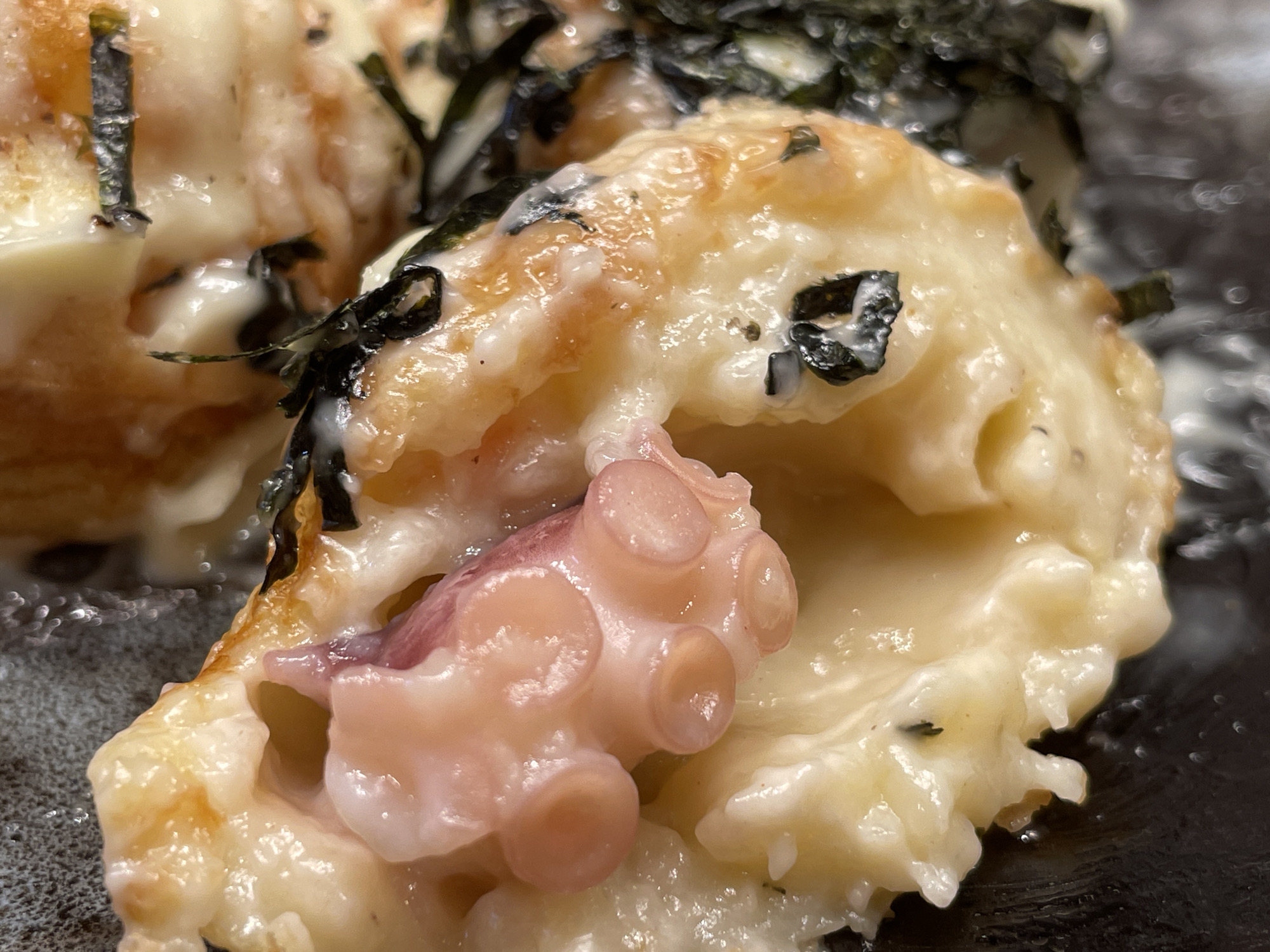 This is a close-up, overhead shot of a takoyaki dish. The takoyaki balls are golden brown with a creamy white sauce drizzled over them and pieces of seaweed scattered on top. One takoyaki ball has been broken open, revealing a pink octopus tentacle inside. The dish is served on a dark, textured plate. The image does not depict any identifiable locations in Taito, Japan.