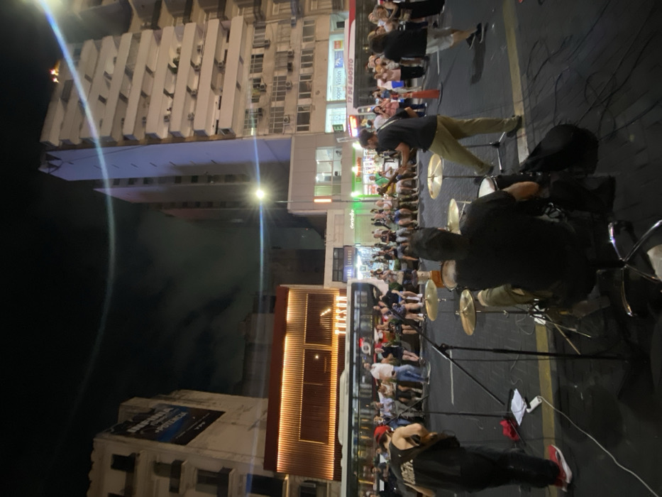 A band performs outdoors at night in a city street. Buildings surround the musicians and a crowd sits on the ground, watching the performance. A drummer is visible in the foreground, along with the tops of his cymbals. Another band member plays guitar while standing further down the street. Streetlights illuminate the scene.