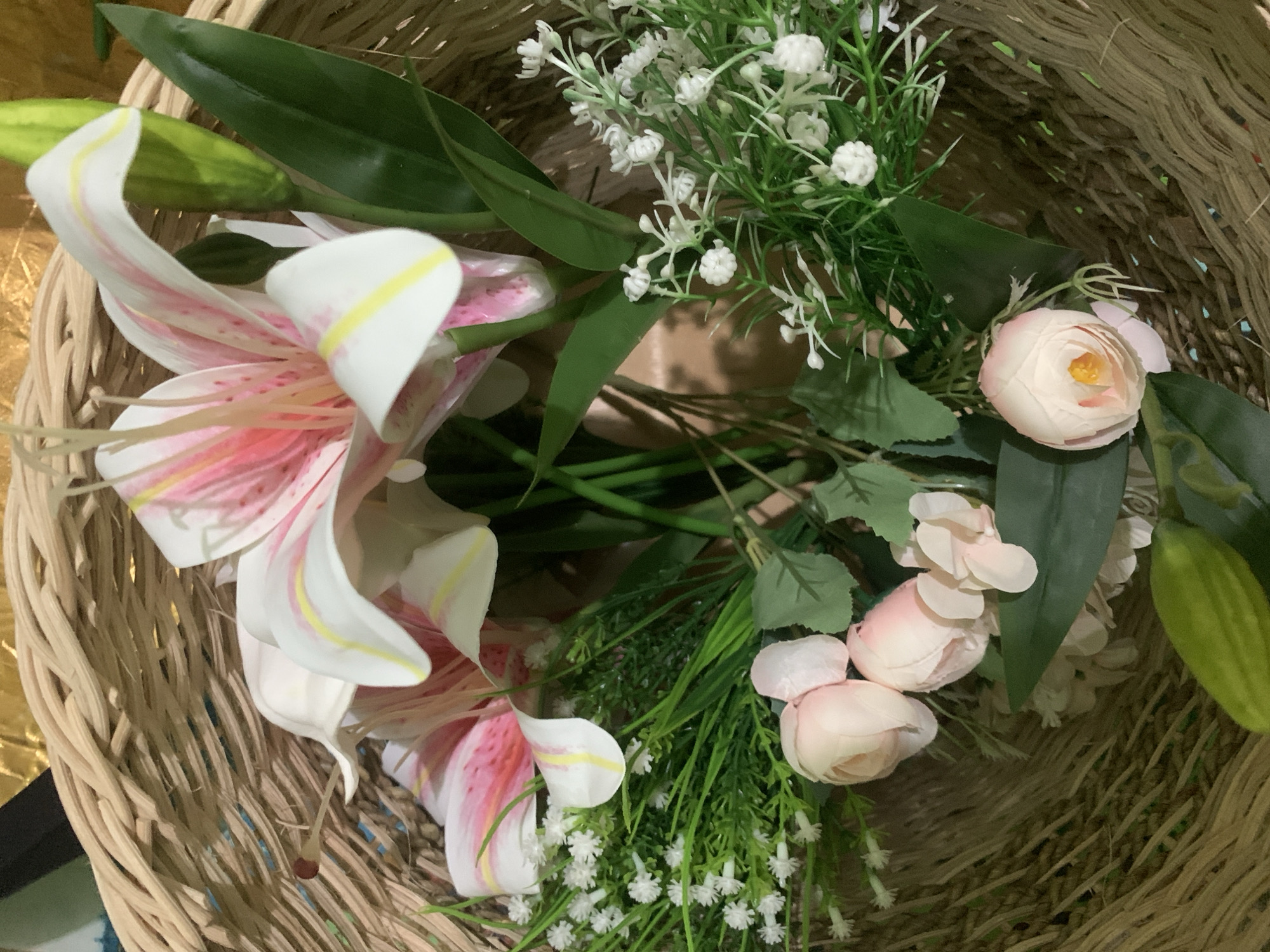 A close-up, overhead shot shows a decorative floral arrangement in a woven basket. The arrangement features large white and pink lilies with yellow accents, interspersed with smaller white flowers and pink roses. Green foliage and buds are also visible. The basket appears to be made of natural fibers and is placed on a surface that is out of focus, but suggests an indoor setting. The lighting is natural and somewhat dim, casting soft shadows on the flowers and basket. The scene is static and no people are present. The image is a still life composition.