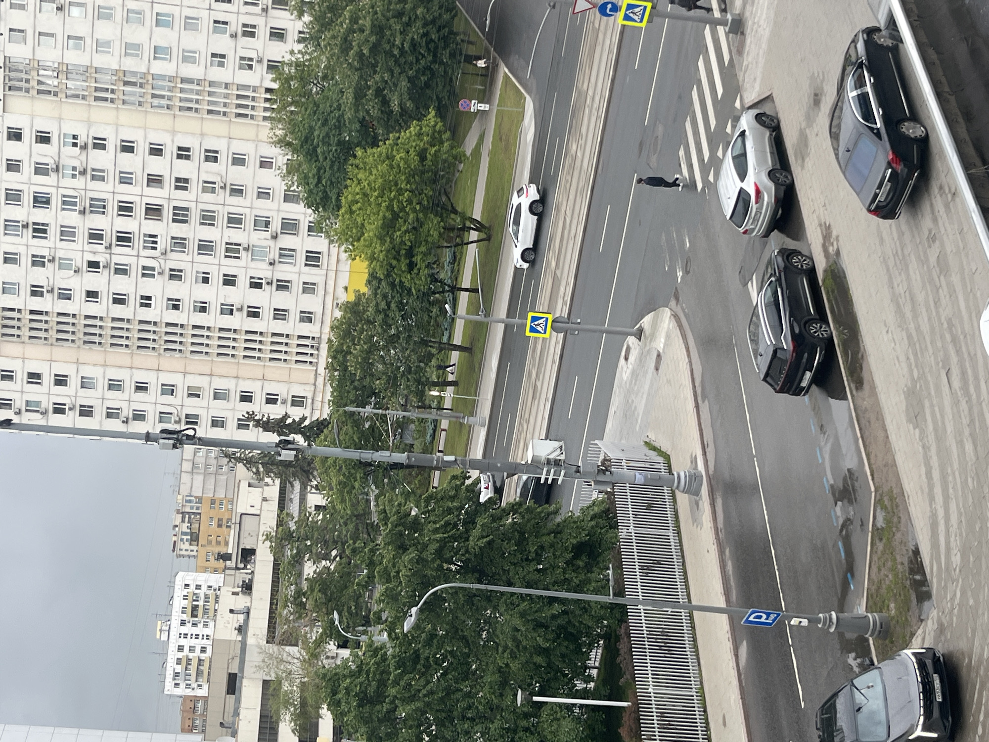 The image shows an urban scene in Moscow, Russia. A large, white building with many windows is visible on the left side. A street runs alongside the building with several parked cars, two black and one white. A pedestrian is crossing the street at a zebra crossing. Streetlights and traffic signs are present. The weather appears overcast. Trees are scattered throughout the scene.