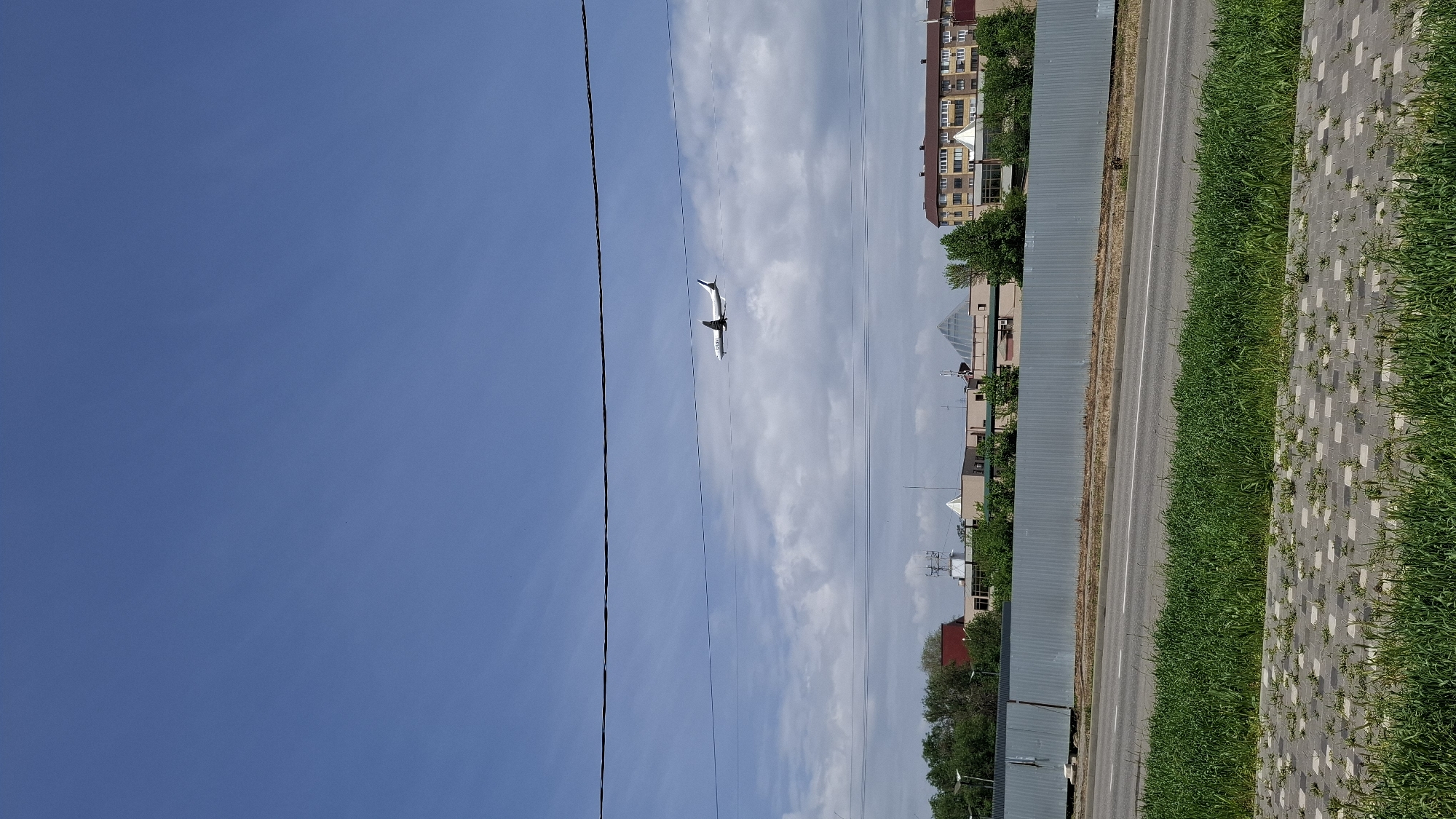 The image captures a clear blue sky with scattered white clouds above a city street in Unknown, Unknown. A passenger airplane is visible in mid-flight. Power lines run parallel to the street. The street features a gray metal fence, a paved road, a green grass strip, and a paved sidewalk. Buildings and trees are visible along the street, suggesting a suburban or residential area.