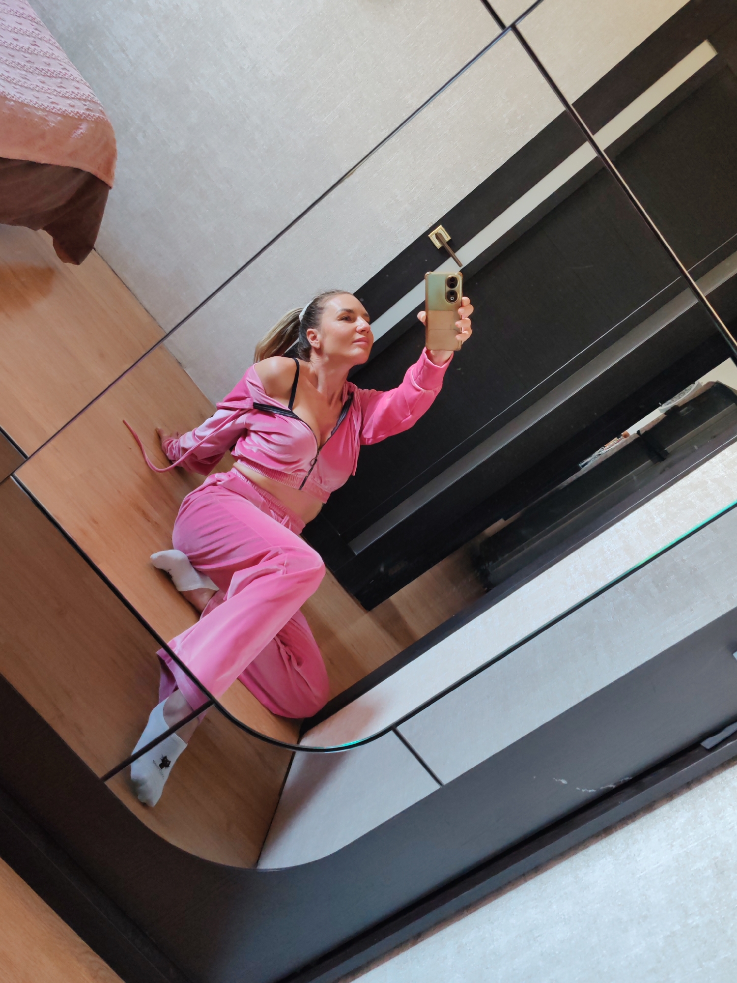 An adult female, with hair pulled back in a ponytail, is depicted taking a mirror selfie in an indoor setting. She is wearing a two-piece, light pink velour tracksuit consisting of a cropped, zippered hoodie and loose-fitting pants. A black bra strap is visible beneath the unzipped top. She wears white ankle socks, the visible left sock displaying a small, dark, stylized emblem. The woman is angled towards the left, appearing to recline or kneel on a light wood-planked floor, holding a light green and tan smartphone vertically in her right hand, aiming it towards her reflection. Her gaze is directed towards the phone/mirror. The scene is reflected in a large, multi-paneled mirror whose segmented design creates prominent diagonal and horizontal lines across the image. Reflections reveal a portion of a bed with a pink, textured blanket and brown base in the upper-left, alongside areas reflecting a light grey, textured wall or ceiling surface and darker, indeterminate elements. A small, light-colored fixture with a dark rod extends from a reflected ceiling in the upper-center.
