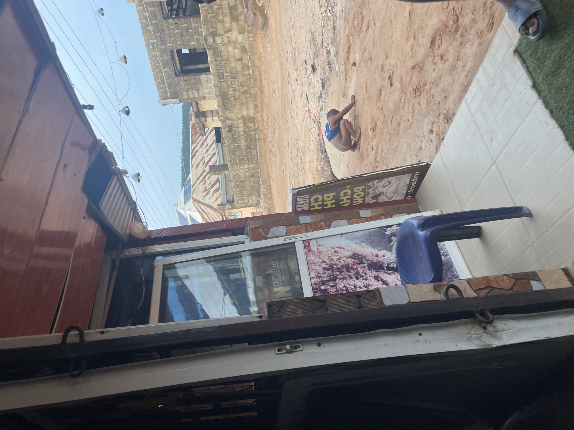 The image depicts an outdoor scene, likely in Lokoja, Nigeria. In the foreground, an open doorway of a shop reveals a tiled floor and a blue plastic chair. A person is crouched on the sandy ground in the mid-ground, appearing to be playing with small objects. Behind this individual, a brick wall with graffiti is visible, and further in the background, buildings with corrugated iron roofs and the sky with suspended lights can be seen. The overall environment suggests a somewhat dusty and undeveloped area.