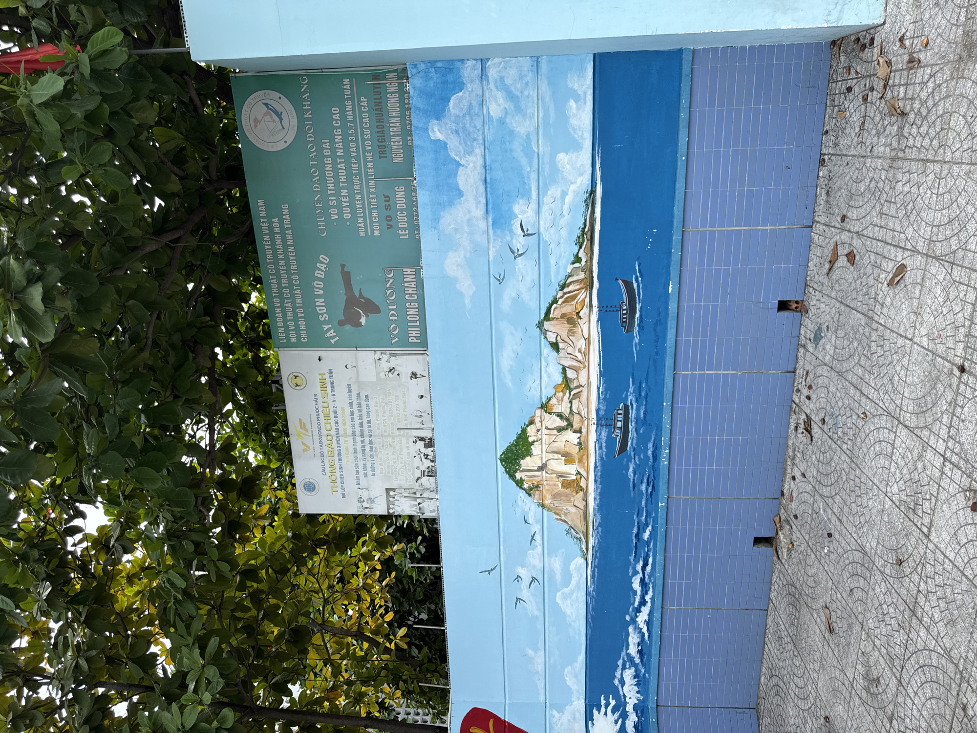 The image captures a section of a blue tiled wall in Nha Trang, Vietnam, adorned with a vibrant mural depicting a coastal landscape. The mural showcases a rocky island with lush green foliage and a sandy beach, set against a bright blue sky filled with fluffy white clouds. The sea below is painted in various shades of blue, with white-capped waves. Two small boats with dark hulls and white superstructures are sailing on the water. Above the mural, a green sign is partially visible, featuring text in Vietnamese related to a martial arts school, identified as "LIÊN ĐOÀN VÕ THUẬT CỔ TRUYỀN VIỆT NAM HỘI VÕ THUẬT KHÁNH HÒA CHI HỘI VÕ THUẬT CỔ TRUYỀN NHA TRANG." The sign advertises classes and training for "TAY SON VO DAO" and "VO DUONG PHI LONG CHANH." A portion of a red flag is also visible at the top right of the image. The foreground shows a gray tiled ground scattered with fallen leaves and debris. A small dark opening is present at the base of the tiled wall.