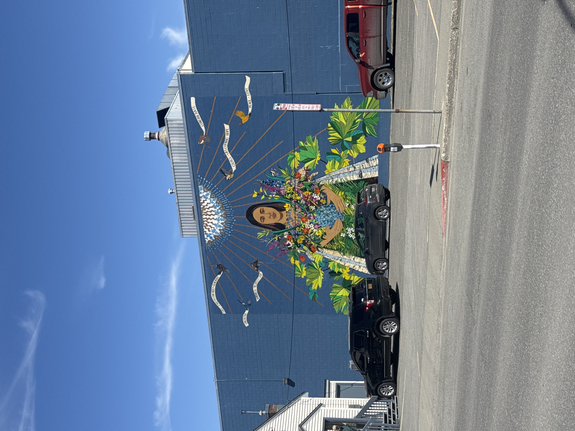 The image shows a large mural on the side of a blue brick building. The mural features a woman holding a bouquet of flowers, with a radiant sun-like design behind her head. There are white banners with words like "CONSIDERATE", "CREATIVE", "GRACE-WEL", and "PEACE-LOVE" written on them, interspersed with images of birds. A red pickup truck is parked next to the building, and two black SUVs are parked in front of the mural. A parking meter and a "No Parking" sign are also visible. A small white building with a metal staircase can be seen at the bottom of the image. The sky is blue with a few wispy clouds. The scene appears to be in a parking lot, with the mural acting as a backdrop. The location is in the city of Anchorage, United States of America.