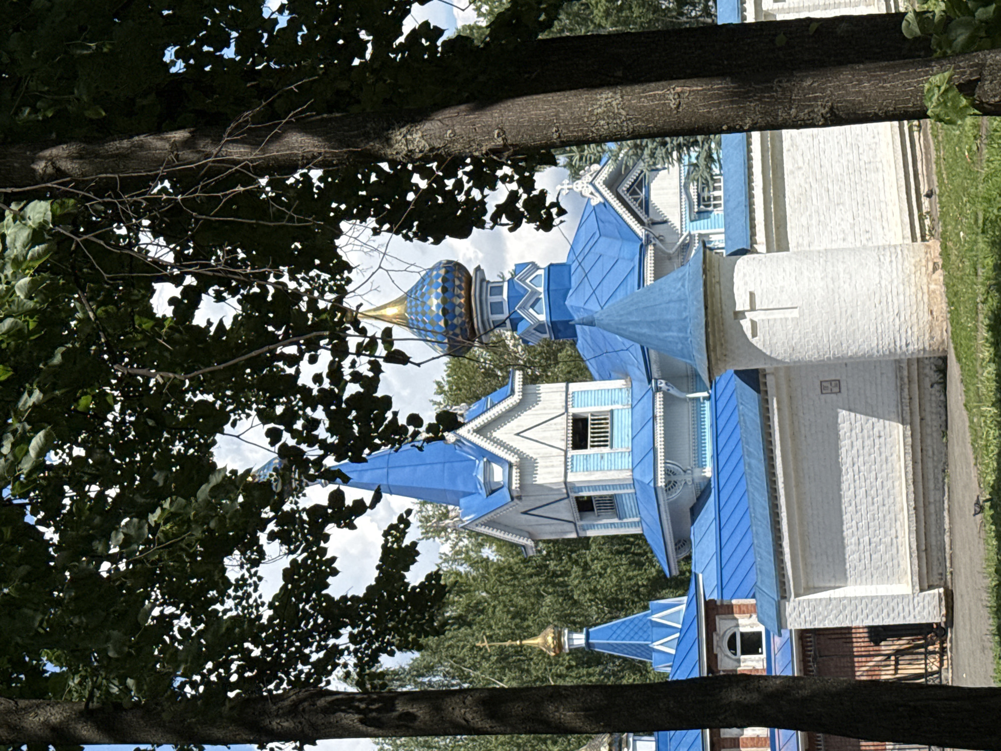 This is a medium shot, eye-level, vertical photograph of a church building, partially obscured by trees in the foreground. The church has a white brick wall with blue roofs and white trim. The central dome is blue with gold accents, and other smaller domes are blue. There is a white turret with a cross on it. The background consists of green trees and a cloudy sky. This is the Church of the Holy Trinity in Izhevsk, Russia. There are no people visible in the image.
