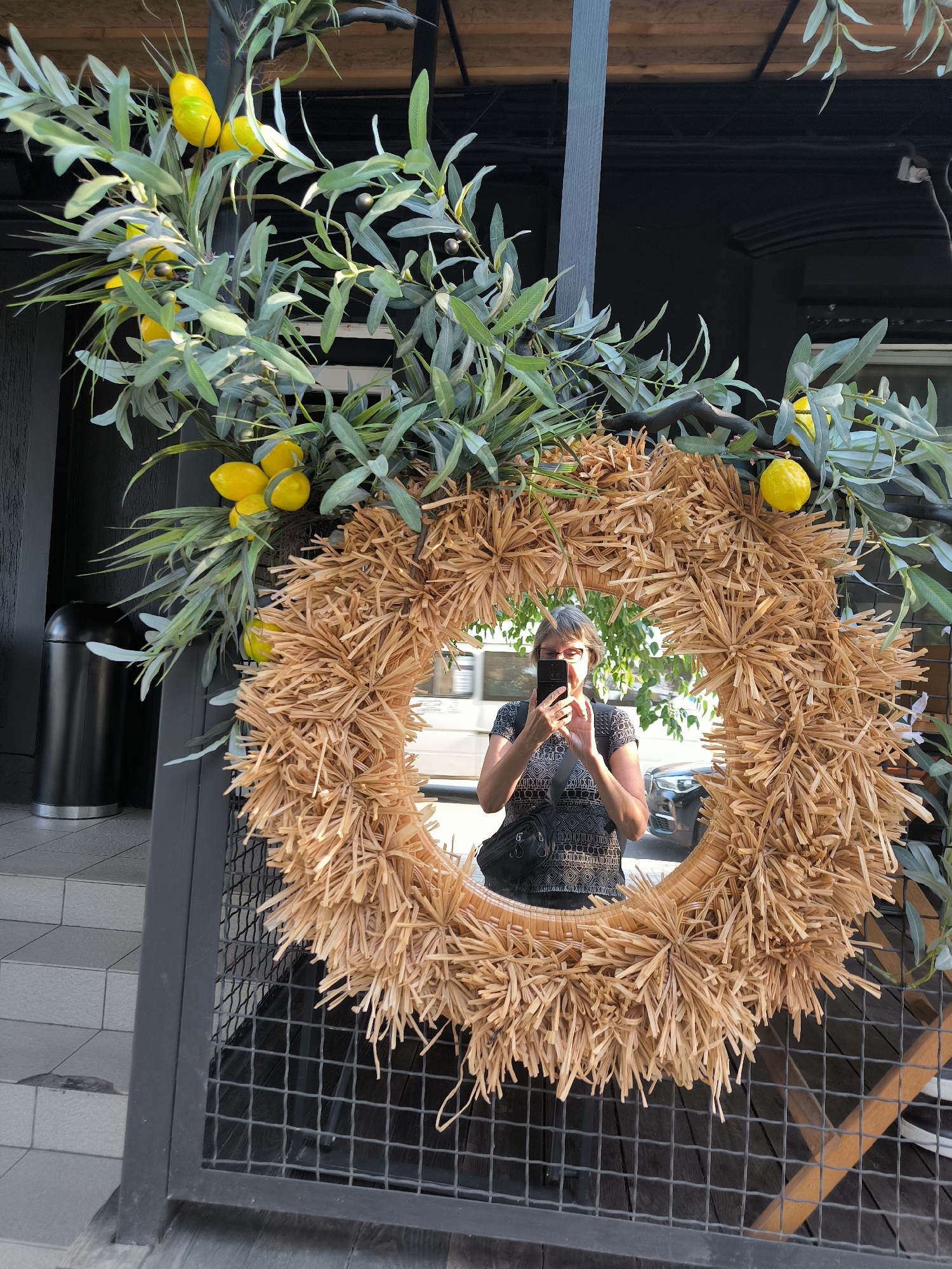 A woman with gray hair and glasses is reflected in a round mirror decorated with a straw wreath and artificial lemon branches. She is wearing a black and white patterned shirt and a black crossbody bag, and is holding a black smartphone up to take the photo. The mirror is surrounded by a frame of straw-like material and green leafy branches with yellow lemons. The background shows a dark building facade and some outdoor seating area with a metal fence. The scene is outdoors, possibly at a cafe or restaurant, with a trash can visible on the left side of the image. The overall impression is a decorative element on a commercial establishment.