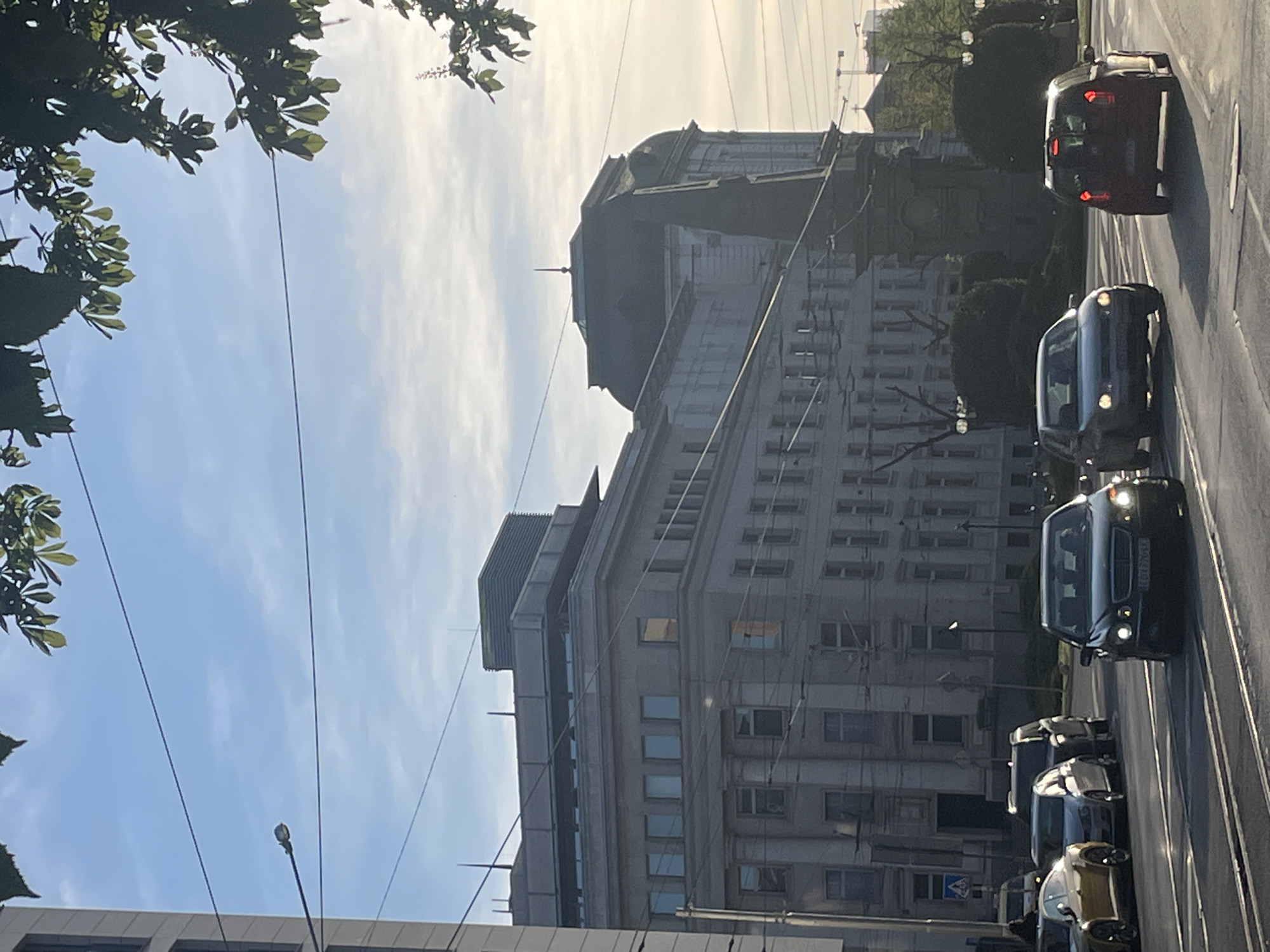 The image shows a street in Sofia, Bulgaria, with several cars driving on the road. A large, light-colored building with a dome is visible in the background. The sky is light blue with some clouds, and trees are visible in the upper left corner of the image. Power lines run across the sky.