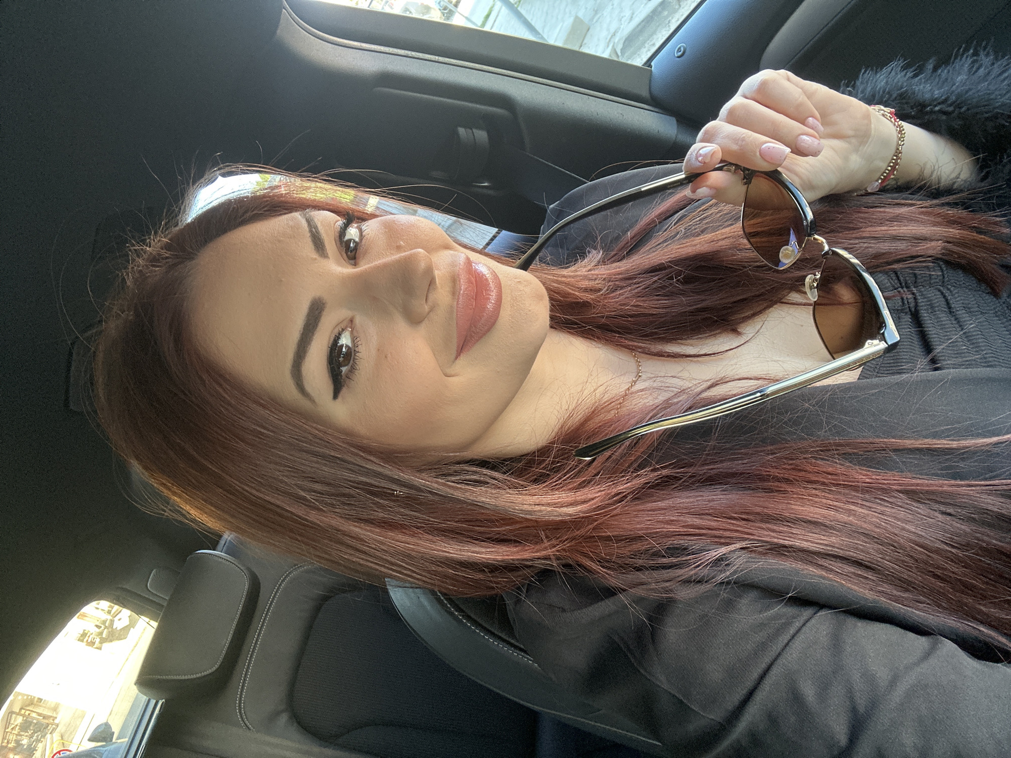 A woman is in the driver's seat of a car. She is holding sunglasses in her right hand. She has red hair. The location is unknown, however, the city is Sofia, Bulgaria. The woman is wearing a black garment.