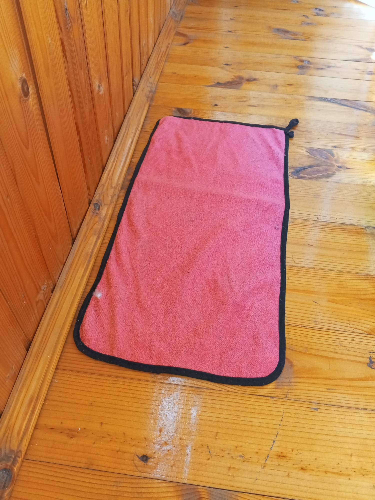 A rectangular pink mat with a black border lies on a wooden floor. The floorboards are oriented horizontally in the frame and are made of light-colored wood with visible grain and knots. To the left of the mat, a vertical wooden wall paneling is visible, also made of light-colored wood. The scene appears to be indoors, possibly a porch or room with wooden construction. The lighting suggests daylight. No people or actions are depicted. The mat has a small black loop attached to one of its corners.