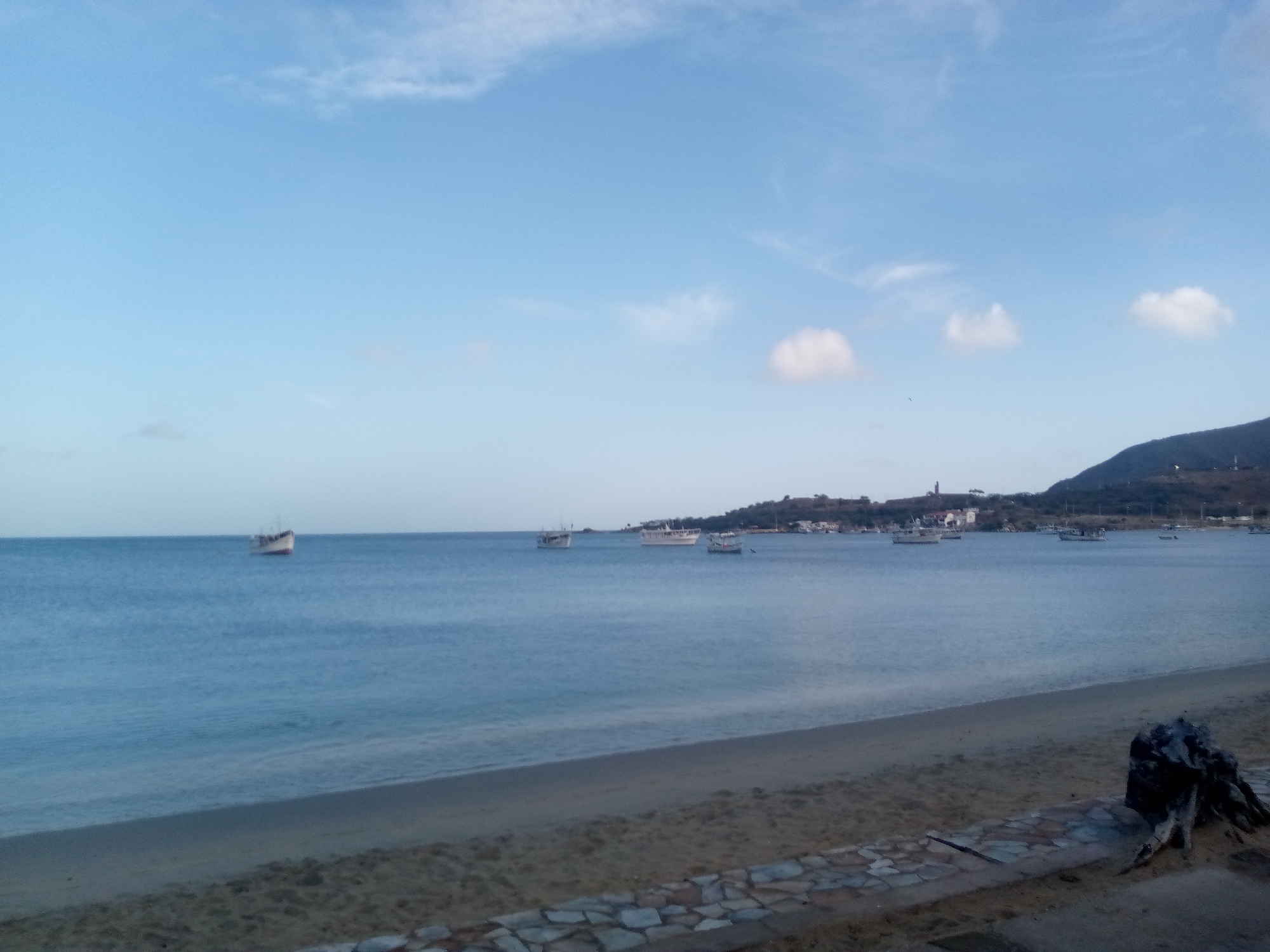 This is a scenic view of a beach and the ocean. The image features a calm sea with several boats anchored in the distance. The shoreline is made up of sand, with a decorative stone border running along the edge of the beach in the foreground. A piece of driftwood is visible on the sand near the stone border.
The sky is a clear, pale blue with a few scattered white clouds, suggesting it is daytime, possibly morning or late afternoon due to the soft light. A hilly landscape with scattered buildings and vegetation forms the background on the far side of the bay, where the boats are moored. The overall mood of the image is peaceful and tranquil. There is no visible text in the image.
