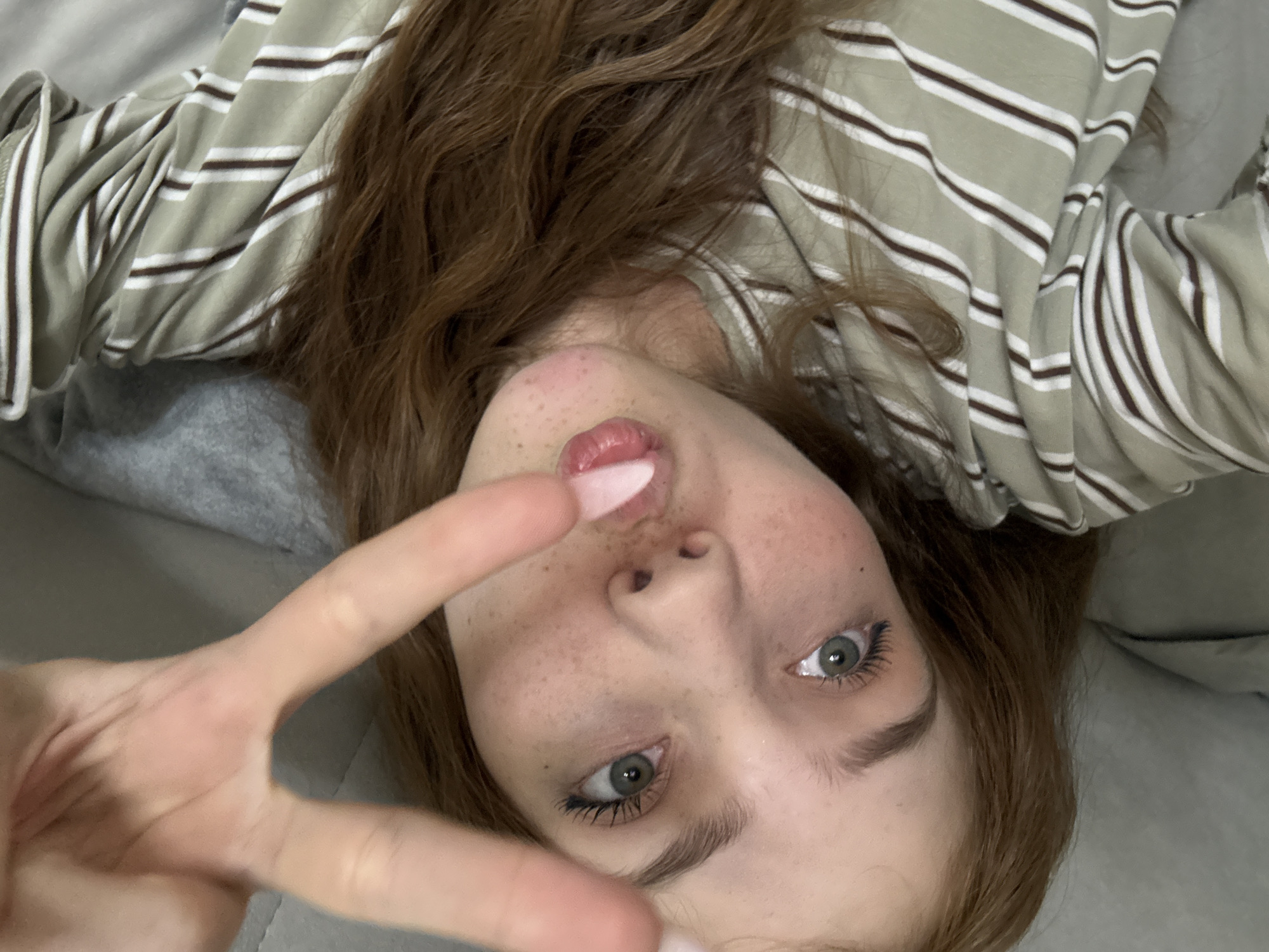 A close-up selfie, taken from an inverted perspective, features a young woman lying down and looking directly into the camera. Her long, wavy reddish-brown hair cascades around her head. She has striking light green or hazel eyes and a scattering of freckles across her nose and cheeks. Her lips are slightly parted, showcasing a glossy finish on her upper lip. She is dressed in a light greenish-grey long-sleeved top adorned with thin white and dark brown horizontal stripes. Prominently in the foreground, her left hand is raised, forming a peace sign, with her nails painted a light pink. The background suggests a soft, grey upholstered surface, likely a bed or sofa in an indoor setting. The expression on her face is neutral yet engaging, with a hint of playfulness. No text is visible in the image. This scene is contextually located in Naberezhnye Chelny, Russia.