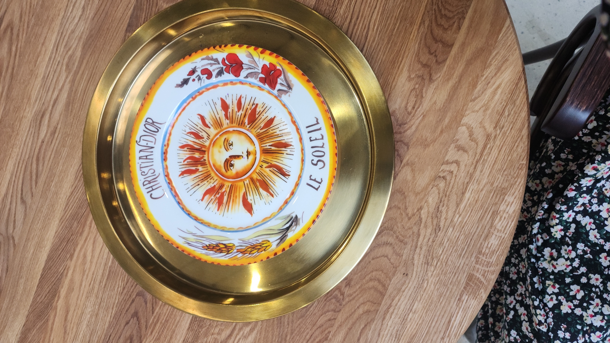 A top-down shot shows a round, brass-colored tray resting on a wooden table. The tray features a decorative plate with a central image of a stylized sun face in orange and yellow tones. The plate also displays the text "CHRISTIAN DIOR" and "LE SOLEIL". Floral and wheat illustrations border the sun image. A portion of a person's floral dress is visible on the right side of the frame. The background appears to be a round wooden table, suggesting an indoor setting. There is no indication of Moscow, Russia.