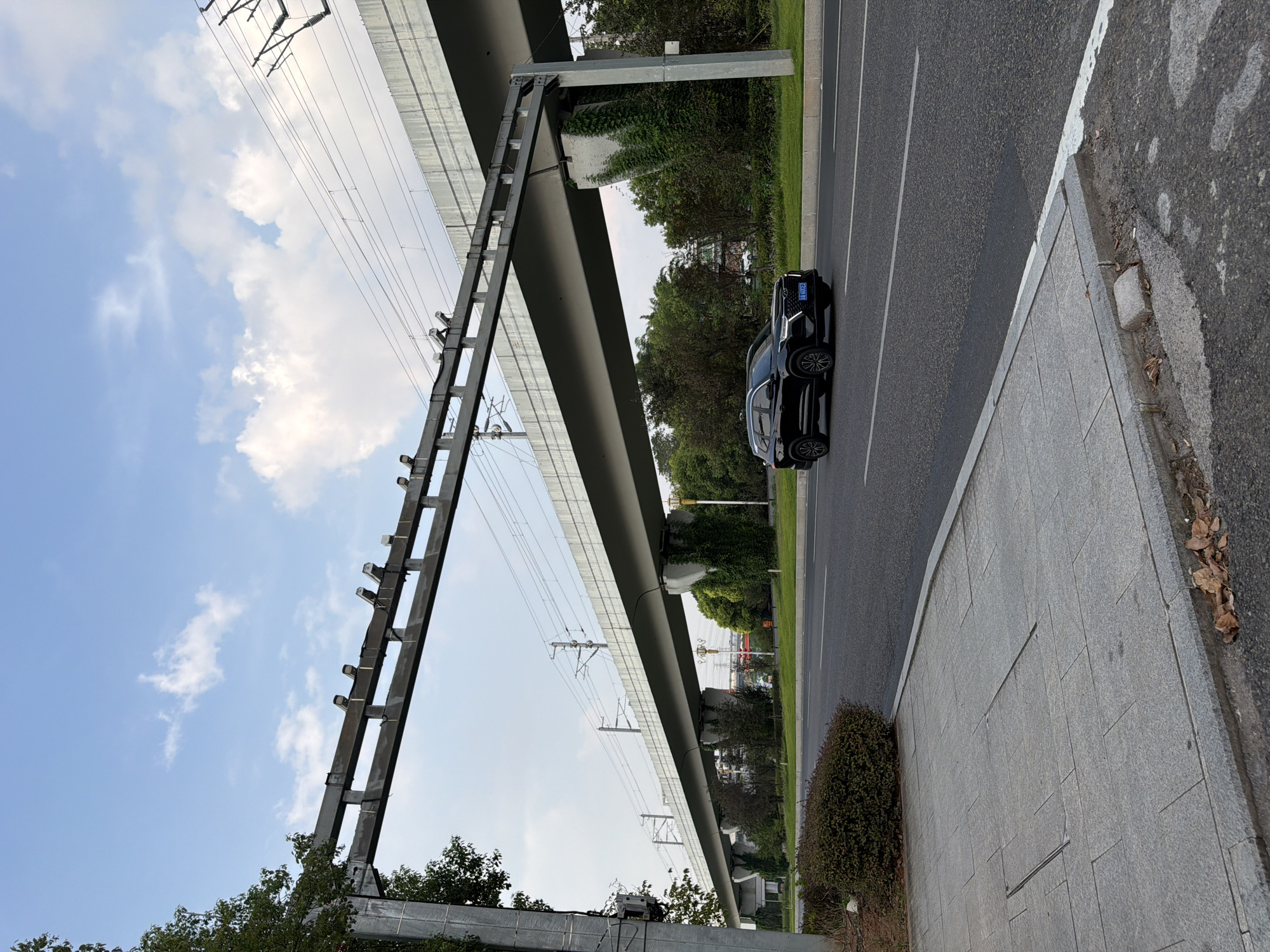 The image depicts a street scene in Jiangjin, China. An elevated railway structure with overhead electrical wires spans across the frame. Beneath the railway, a black SUV drives on a paved road. The road has lane markings and a sidewalk made of grey paving stones is visible in the foreground. Green vegetation, including bushes and trees, lines the side of the road and covers some of the railway supports. The sky above is blue with scattered white clouds. A metal frame with what appear to be lights or sensors is suspended from a pole on the left side of the image.