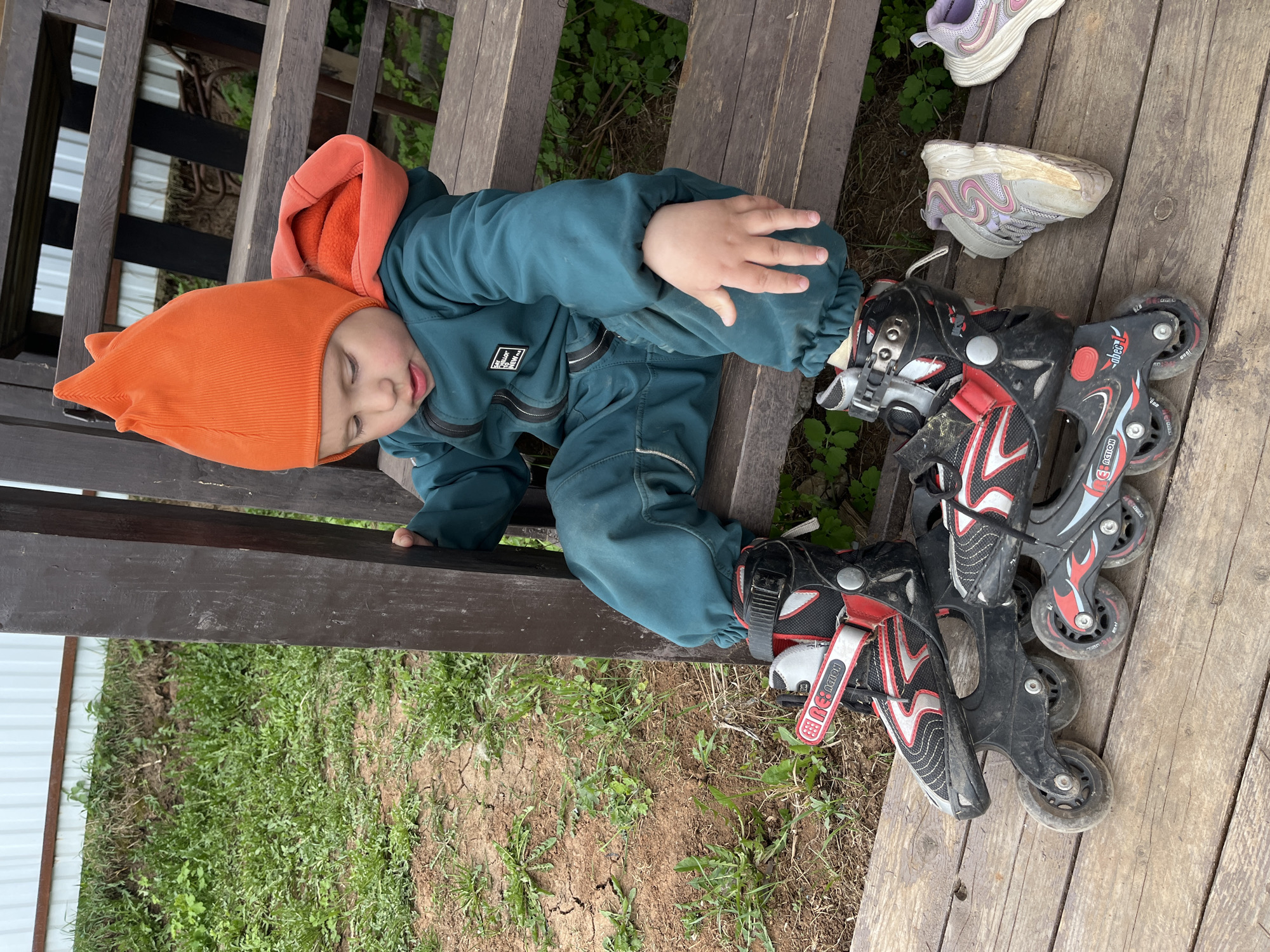A young child, wearing a teal jumpsuit with an orange hood and matching pointed hat, is seated on a weathered wooden staircase or platform. The child is wearing roller skates and appears to be either resting or struggling with the skates. Beside the child are a pair of older, worn-out athletic shoes. The background consists of patchy grass, dirt, and a glimpse of what appears to be a white fence or wall, suggesting an outdoor setting near a building. The image was taken in Chaikovsky, Russia.