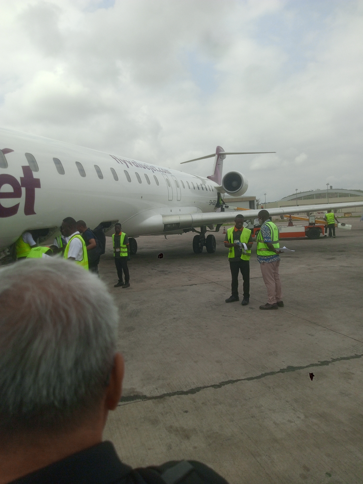 The image shows a white commercial jet, possibly a Bombardier CRJ, parked on an airport tarmac in Gwaska, Nigeria. The tail fin is marked with purple. Ground crew wearing reflective vests are present near the plane, some appearing to load or unload cargo, while others stand and converse. The plane's registration number, "5N-CEX", is visible near the rear. In the foreground, the back of a person's head is visible. The sky is overcast. An airport control tower can be seen in the background. There is a small red blotch on the tarmac near the lower right corner.