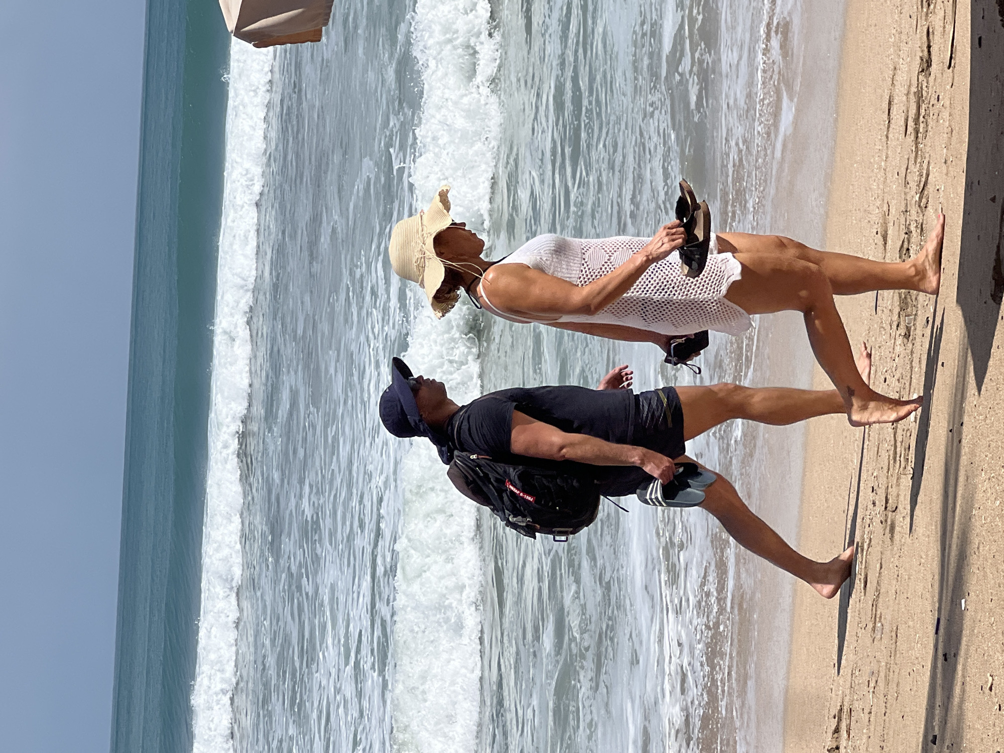Two individuals, a man and a woman, walk side-by-side along the sandy shore of Kuta Beach, Indonesia. The man, wearing a black t-shirt, dark shorts, and a dark blue baseball cap, carries a black backpack. He is holding a pair of black and white flip-flops in his right hand. The woman, dressed in a white cover-up dress and a wide-brimmed straw hat, walks to his right. She holds a pair of brown sandals in her left hand. Both are facing the same direction, walking towards the right side of the frame. The ocean, with gentle waves breaking on the shore, forms the backdrop. The sky is clear and blue, suggesting a sunny day. The sand is a light brown color, with some scattered debris visible. The overall scene depicts a relaxed stroll on a beach.