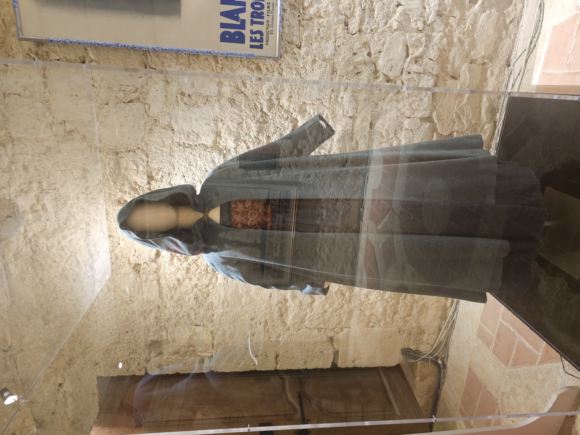 The image shows a mannequin dressed in a historical costume, displayed behind glass in what appears to be a museum or exhibition space. The costume consists of a long, hooded, grey cloak over a patterned dress. The background is a rough stone wall, and to the left, there is a wooden door with ornate carvings. A poster with French text is visible on the right side of the wall. The floor is tiled with terracotta-colored squares. The scene is lit by overhead spotlights. This display is located in the Château de Lavardens, in Lavardens, France.