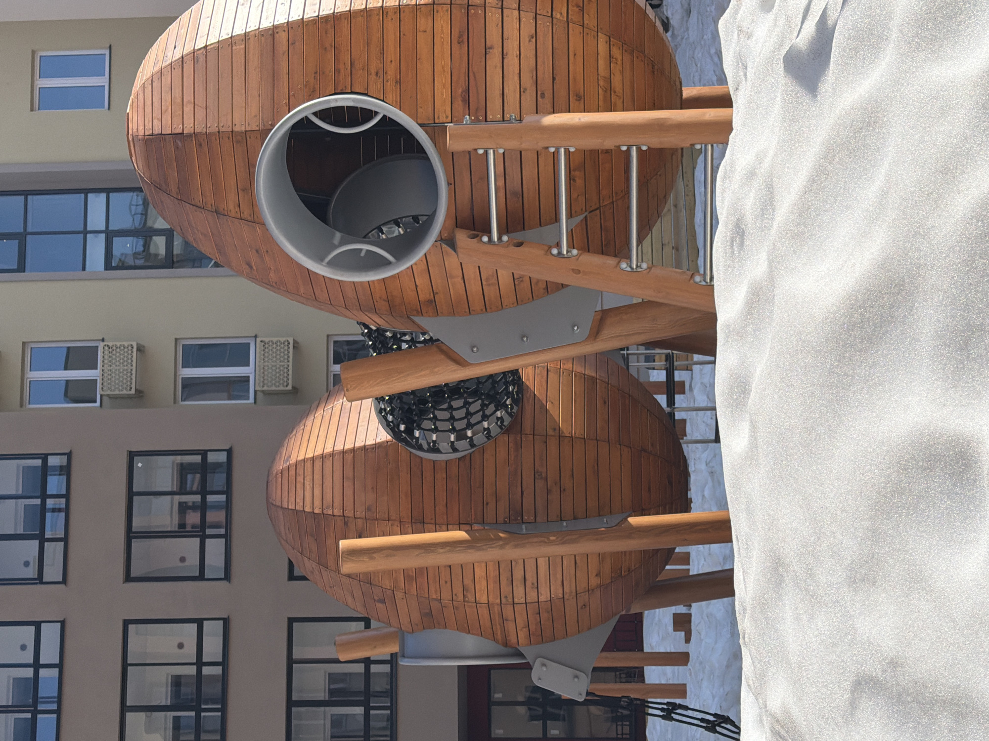 The image shows a playground structure resembling two wooden airships connected by a spiral staircase. The airships are made of narrow vertical wooden planks and feature circular openings with metal framing. One opening has a gray metal tube leading into it. The staircase is constructed with wooden rails and metal supports. A large, light-colored, soft-looking structure partially obscures the right side of the airships. In the background, there is a multi-story building with numerous windows and air conditioning units visible on the upper floors. The scene is located in Pervomaysky, Russia.