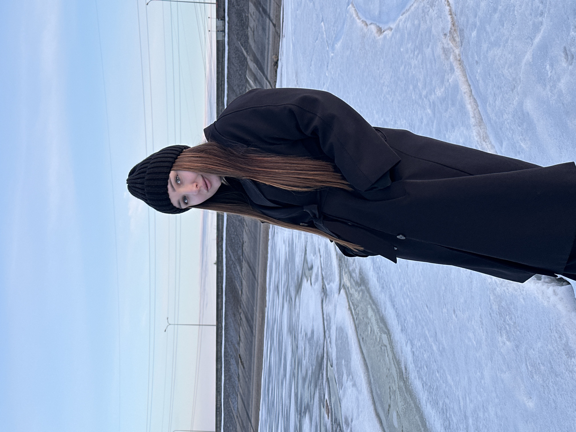 A young woman is standing near the embankment of the Kama River in the city of Chaikovsky, Russia. She is wearing a black coat and a black knitted hat. She is turned toward the camera. The river is partially frozen. The sky is pale blue. A concrete barrier and lampposts are visible in the background.