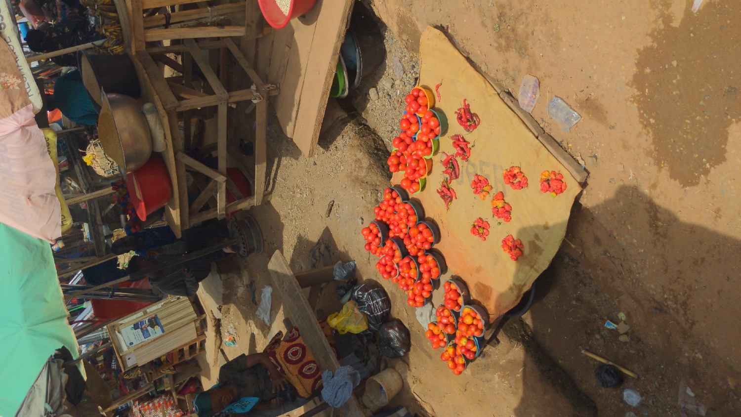 This image captures an outdoor market scene in Ado, Nigeria, viewed from a slightly elevated perspective. The ground is uneven, dusty earth, characteristic of an informal open-air market. The main focus is on a vendor's display of fresh produce arranged on a light-colored tarp or sheet spread directly on the ground.
Numerous small, red tomatoes are meticulously portioned into individual colorful (blue, green, black) plastic bowls, creating neat rows across the tarp. Adjacent to the tomatoes, several piles of bright red and orange peppers, likely scotch bonnets, are also displayed for sale.
To the left of the produce display, a makeshift wooden stall or stand is visible, constructed from rough planks and beams. Within and around this structure, various household items and market wares are scattered, including large metal basins, a red plastic bowl filled with what appears to be grains, and other containers. A light green umbrella or canopy edge is visible on the far left, suggesting protection from the sun.
Scattered on the ground are black and yellow plastic bags, as well as some discarded plastic wrappers and debris, indicating active market commerce. Part of a person's leg and a hand are visible in the lower-left corner, clad in patterned fabric, suggesting a vendor or customer browsing.
The scene is brightly lit by the sun, casting distinct shadows and suggesting it is daytime. The overall impression is one of a bustling, functional market environment. Faint, dark text, possibly "FOOD," is visible on the tarp near the peppers. Further back on a wooden structure, a white poster with indiscernible text and images is affixed.