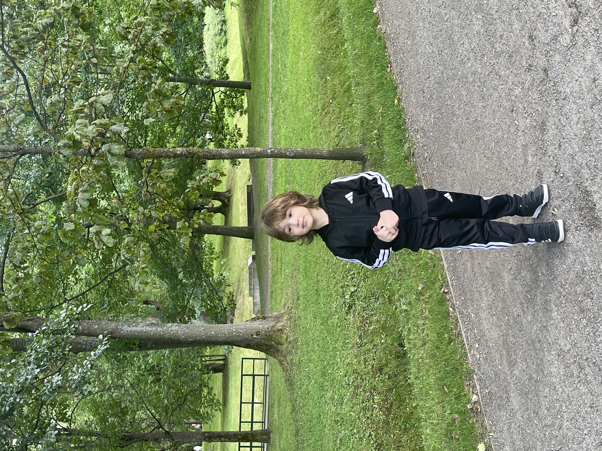 A young boy stands on a paved pathway in a park. He is wearing a black tracksuit with white stripes and black sneakers. The boy has medium-length, wavy brown hair and is looking slightly to his left. Behind him, the park features lush green grass, trees with dense foliage, and some concrete structures. A black metal fence is visible in the background to the left. The overall lighting suggests an overcast day.