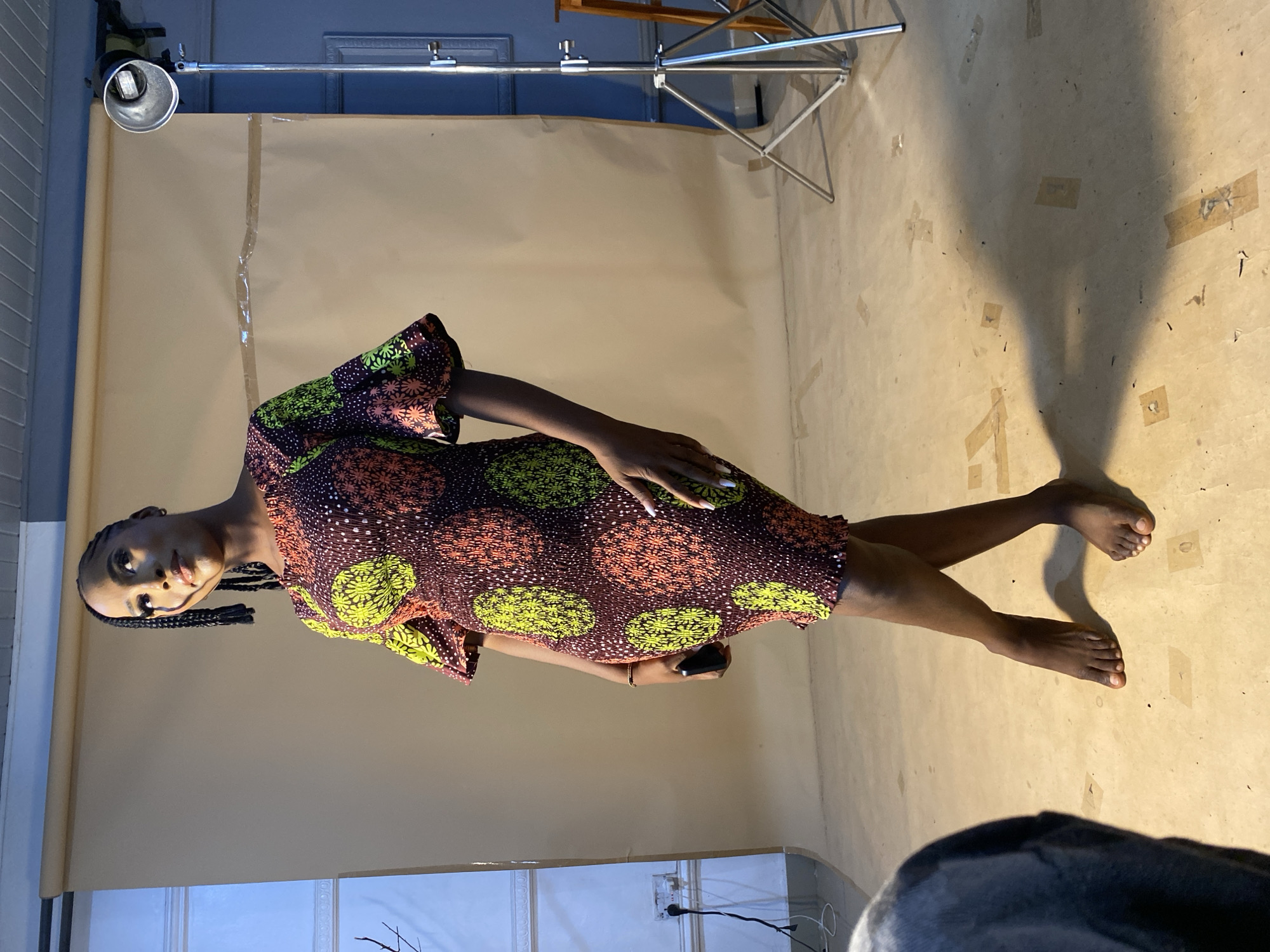 The image shows a young Black woman standing in what appears to be a studio setting in Bukuru, Nigeria. She is wearing a short, colorful dress with a brown base and a pattern of lime green and orange circles and star-like shapes. The dress has bell sleeves and a gathered or ruched texture. Her hair is styled in braids that fall over her shoulders, and she is looking upwards and to her left with a slight smile. Her left hand is holding a dark-colored mobile phone, and her right arm is resting at her side. Her legs are crossed at the knees, and she is barefoot.
The background consists of a large, plain, light brown or tan backdrop, likely made of fabric or paper, stretched taut. To the right of the woman, a professional lighting stand with a silver reflector is visible. In the foreground, the bottom left corner of the image shows a portion of a dark-colored object, possibly a chair or part of a person, and a shadow cast on the floor. The floor itself is a light tan color and appears to be a seamless surface, with some visible pieces of tape scattered across it. The overall lighting suggests a controlled studio environment.