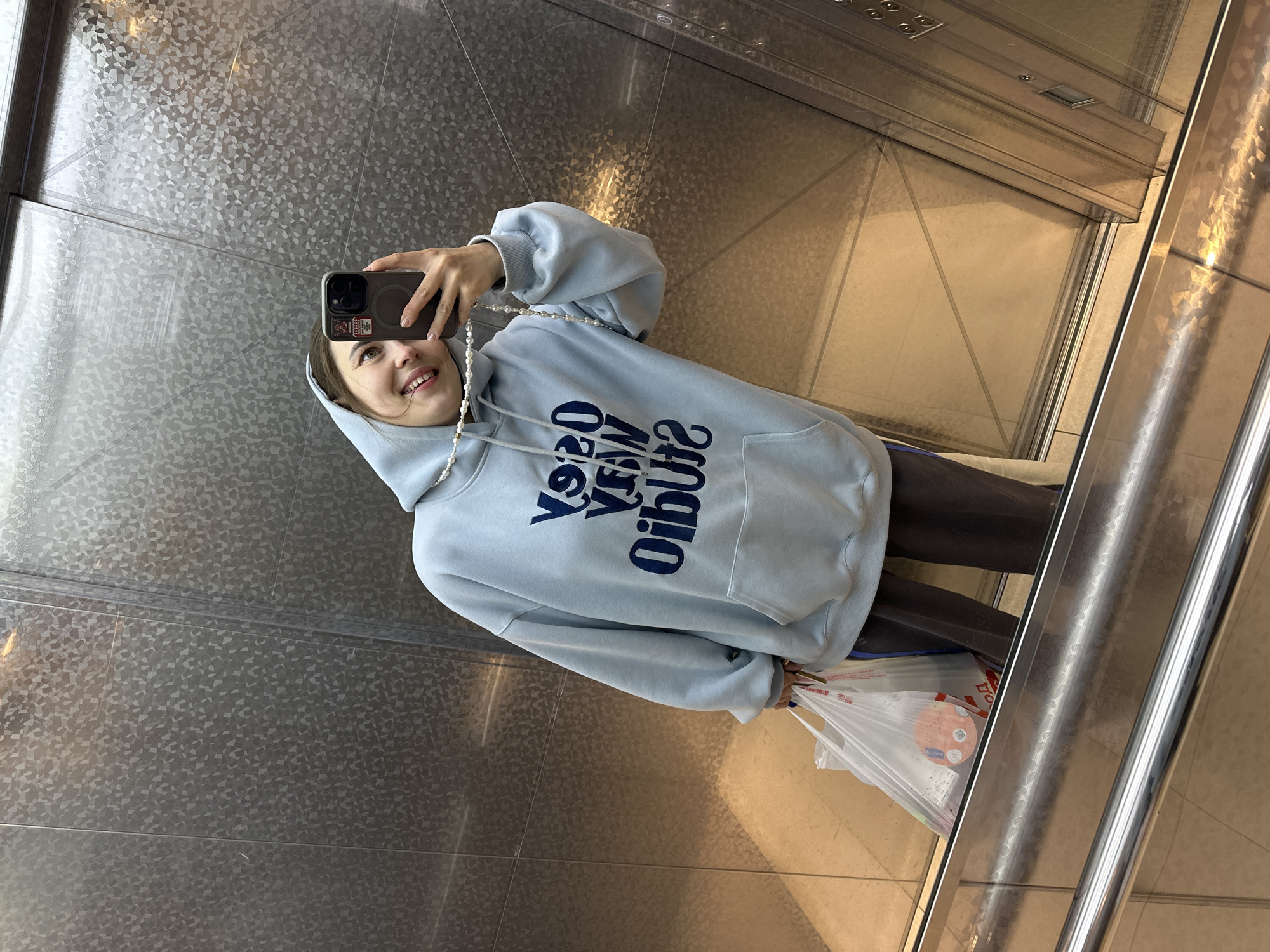 A young woman is depicted taking a selfie in a reflective surface, likely an elevator mirror or polished wall, in Perm, Russia. She smiles broadly at the camera, holding a dark-cased smartphone in her right hand, which has a small sticker on its back and a pearl-beaded strap attached. She is dressed in a light blue oversized hoodie, featuring dark blue, mirrored text across the chest that appears to read "YOYO VERY STUDIO". In her left hand, she carries a white plastic shopping bag adorned with red lettering and a circular graphic, suggesting recent purchases. The background reveals the interior of a modern space, possibly an elevator, with patterned, metallic silver walls and a light-colored tiled floor. Reflections of the ceiling with light fixtures and a shiny chrome handrail are also visible. Her cheerful expression suggests a casual and positive mood during her outing.