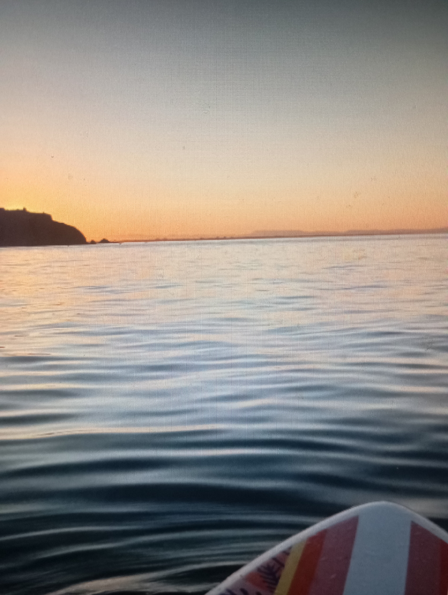 The image depicts a serene body of water at sunset. The sky is a gradient of soft oranges and blues, with a silhouetted coastline visible in the distance. Gentle waves ripple across the water's surface, reflecting the colors of the sky. In the foreground, the edge of a surfboard with a colorful striped pattern is visible, suggesting someone is on the water. The overall scene evokes a peaceful and tranquil atmosphere. The location is not specified in the image, and therefore cannot be determined.