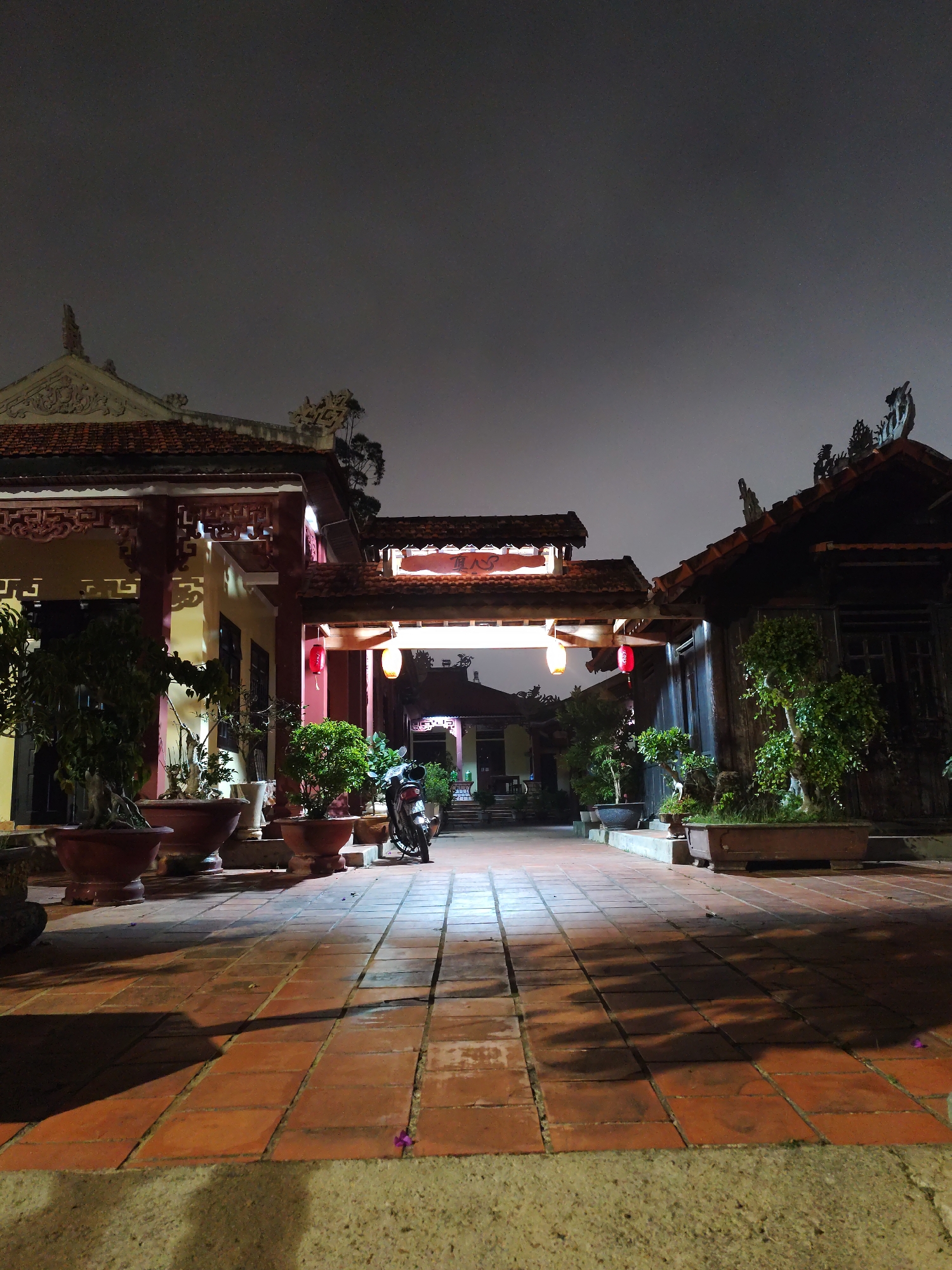 The scene depicts a courtyard with traditional Vietnamese architecture, possibly in Da Lat, Vietnam, based on the style. The courtyard is paved with reddish-brown bricks. Two buildings flank the sides of the courtyard, connected by an ornate gateway adorned with hanging red lanterns. The buildings have sloping tiled roofs with decorative features. Potted plants and bonsai trees are placed throughout the courtyard, adding to the aesthetic. A motorcycle is parked in the center of the courtyard, its headlight illuminating the brick path. The sky is overcast, and it appears to be night. The overall lighting is dim, with light sources from the lanterns and the motorcycle headlight.