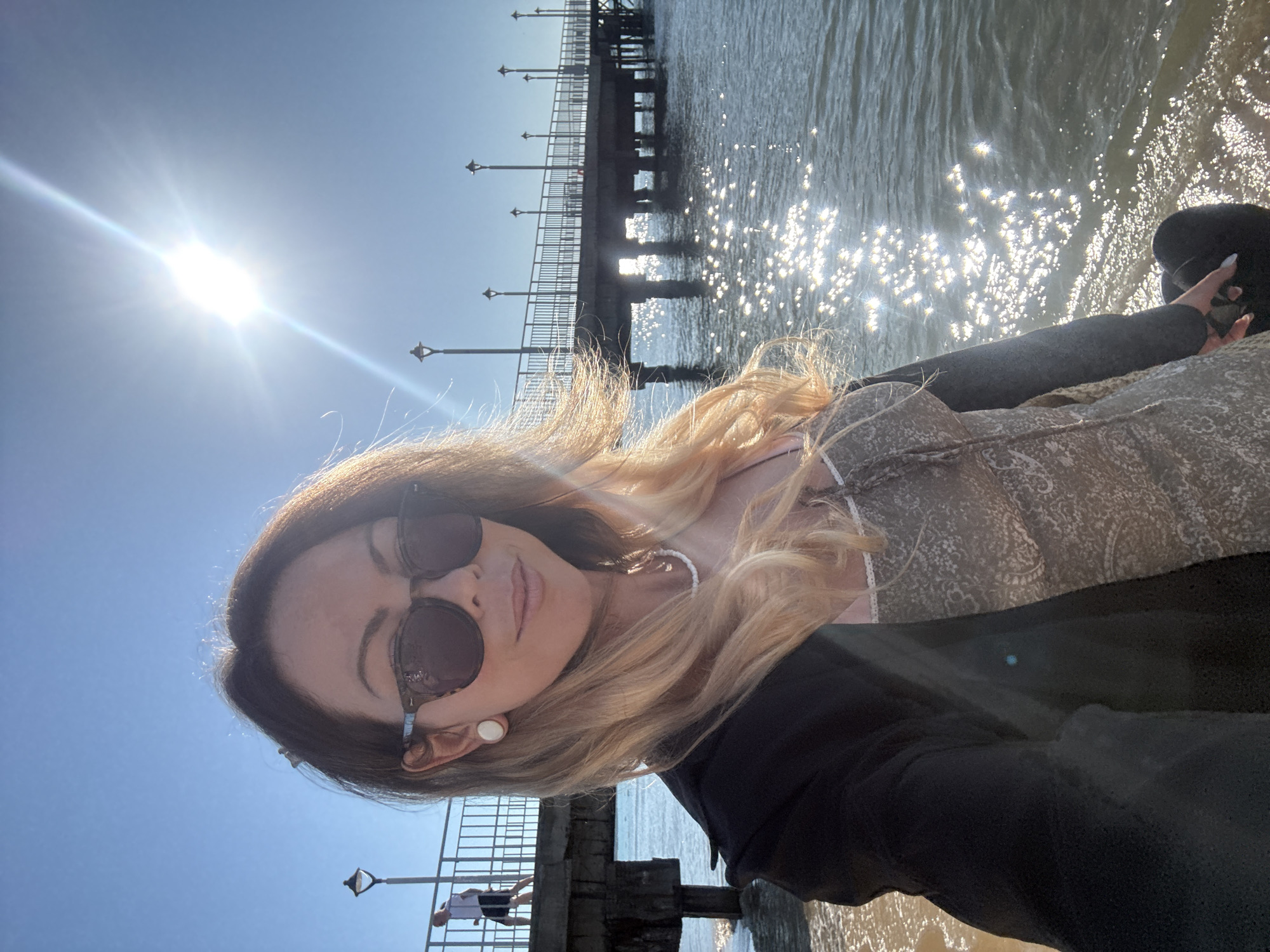 A woman is taking a selfie on a beach in Nesebar, Bulgaria, with a pier in the background. The sun is shining brightly in the clear blue sky, reflecting off the water. The woman is wearing sunglasses, a light-colored top with a patterned overlay, and a dark jacket. She has blonde hair and is wearing large white earrings. In the background, a person is walking on the pier, which has a railing and lampposts. The water is calm with sunlight glinting on its surface.
