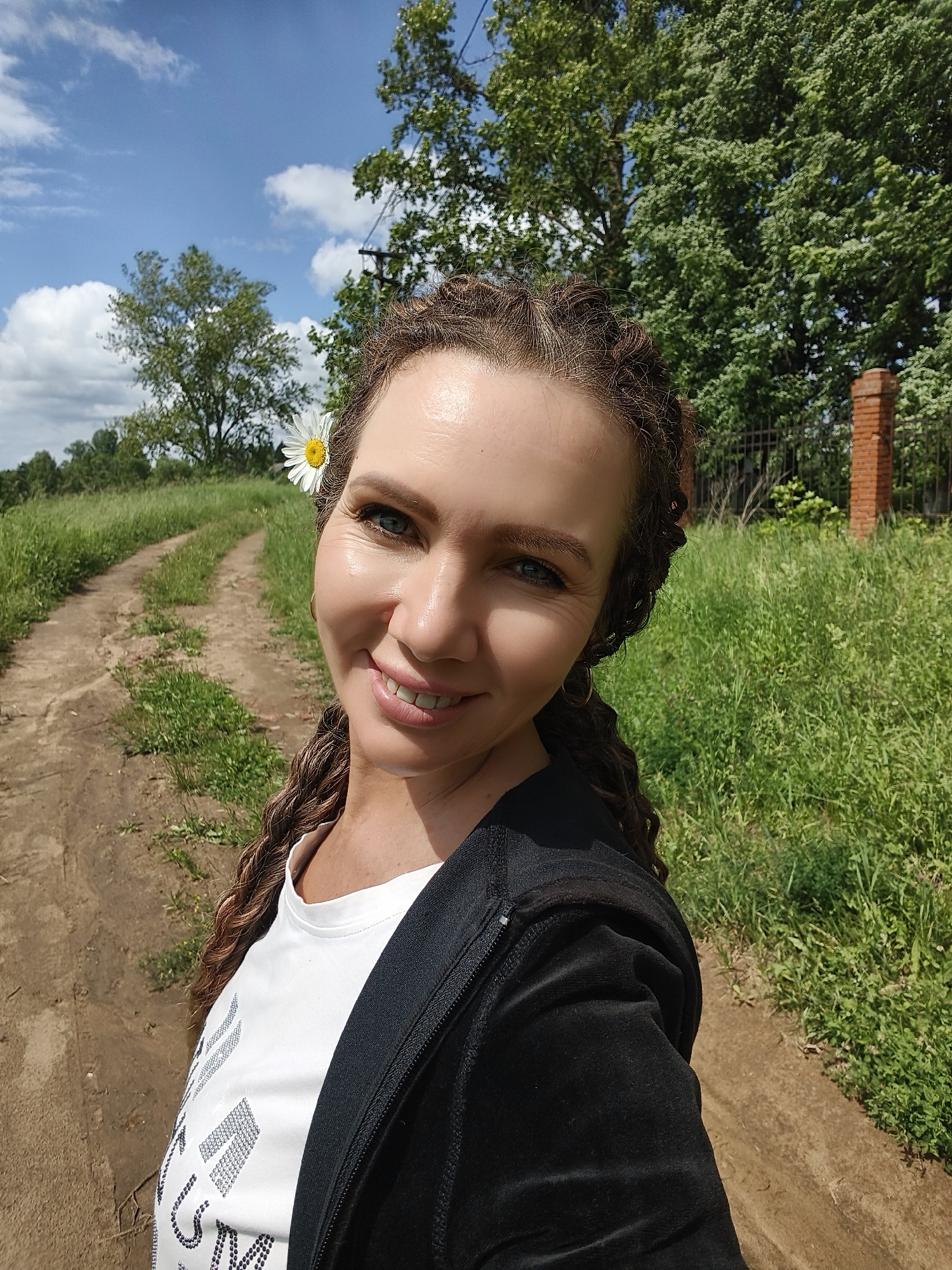 A woman with braided brown hair and a daisy tucked behind her ear smiles at the camera, holding the camera out to take a selfie. She is wearing a white t-shirt with a sparkly design and a black zip-up hoodie. The background is a dirt road lined with tall green grass. In the distance, there are trees, a brick pillar, and a fence. The sky is blue with scattered white clouds. The image appears to have been taken in Zavod Mikhailovskii, Russia.