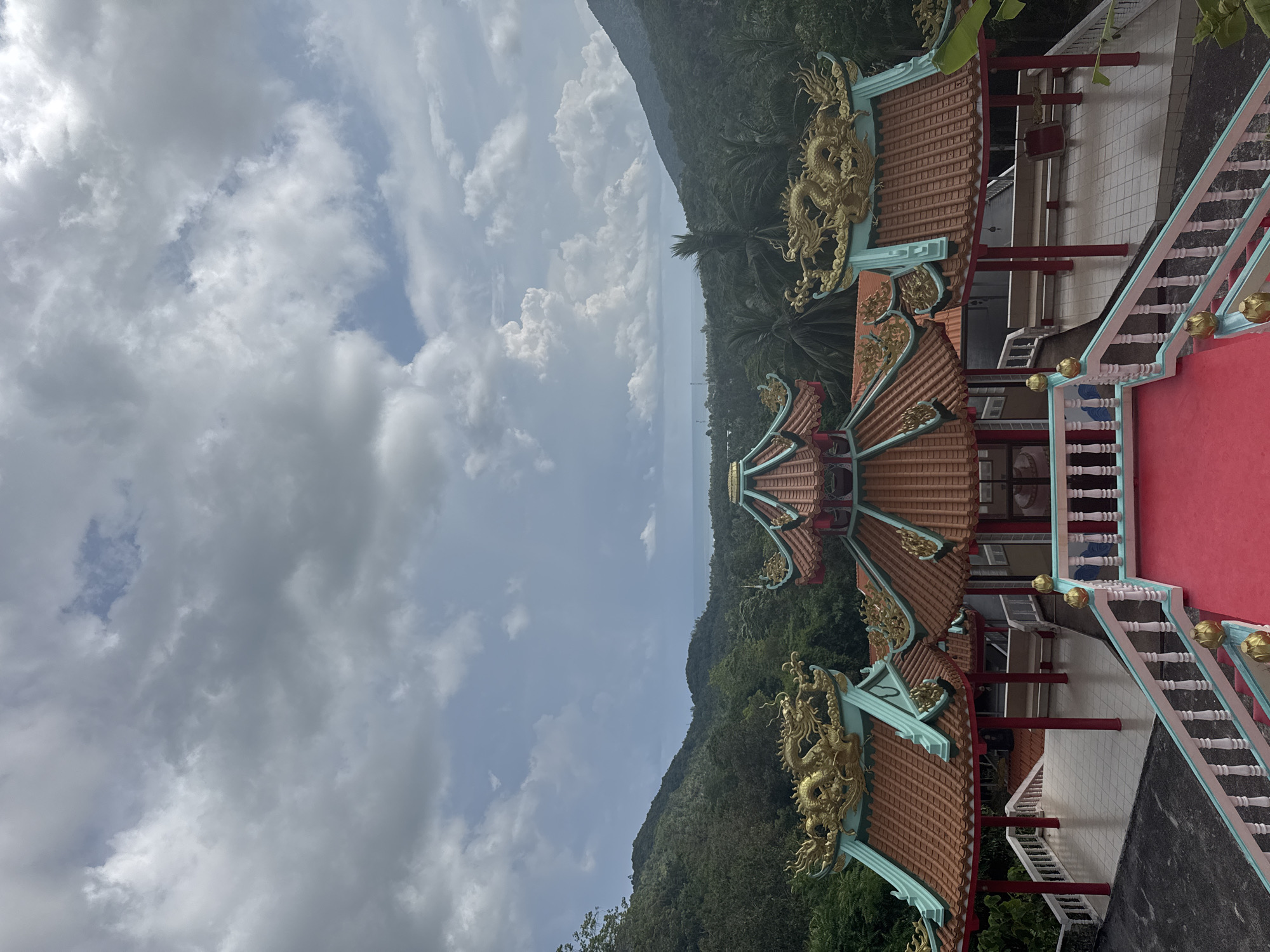 The image shows a temple structure in Moo 7, Thailand. The temple has multiple levels with red tiled roofs adorned with golden dragon sculptures. The roofs are supported by turquoise-colored eaves. Red pillars and a red carpet are visible on the temple's upper levels. The temple is situated on a hillside, with lush green vegetation surrounding it. In the distance, the sea is visible, along with a cloudy sky. There are no people in the scene.