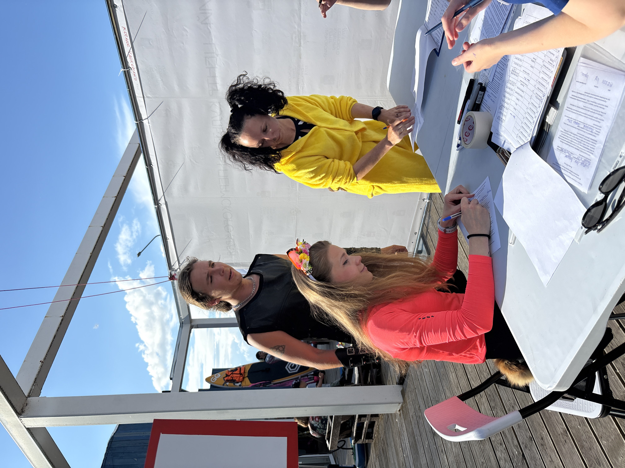 The media file shows an outdoor scene at what appears to be an event or registration area. Three people are visible at a table. On the left, a man with long blonde hair and a black t-shirt looks towards the right. Next to him, a woman with dark hair pulled into pigtails wears a bright yellow robe or jacket and is writing on a document. To the right, a woman with long blonde hair and a floral headband is wearing a vibrant orange-pink long-sleeved shirt and is also writing on a document. The table is covered with papers, pens, tape, and sunglasses, suggesting administrative tasks are being performed. In the background, there is a white tent or awning and a clear blue sky with some clouds. Wooden planks are visible below the table, indicating an outdoor deck or pier. A surfboard with the word "GO" is leaning against a structure in the background. The lighting suggests it is daytime, likely sunny. There is no readily discernible text in the visible portion of the image, other than the word "GO" on the surfboard. The overall mood appears to be active and possibly related to a competition or organized activity. The location context suggests this event is taking place in Chaikovsky, Russia.