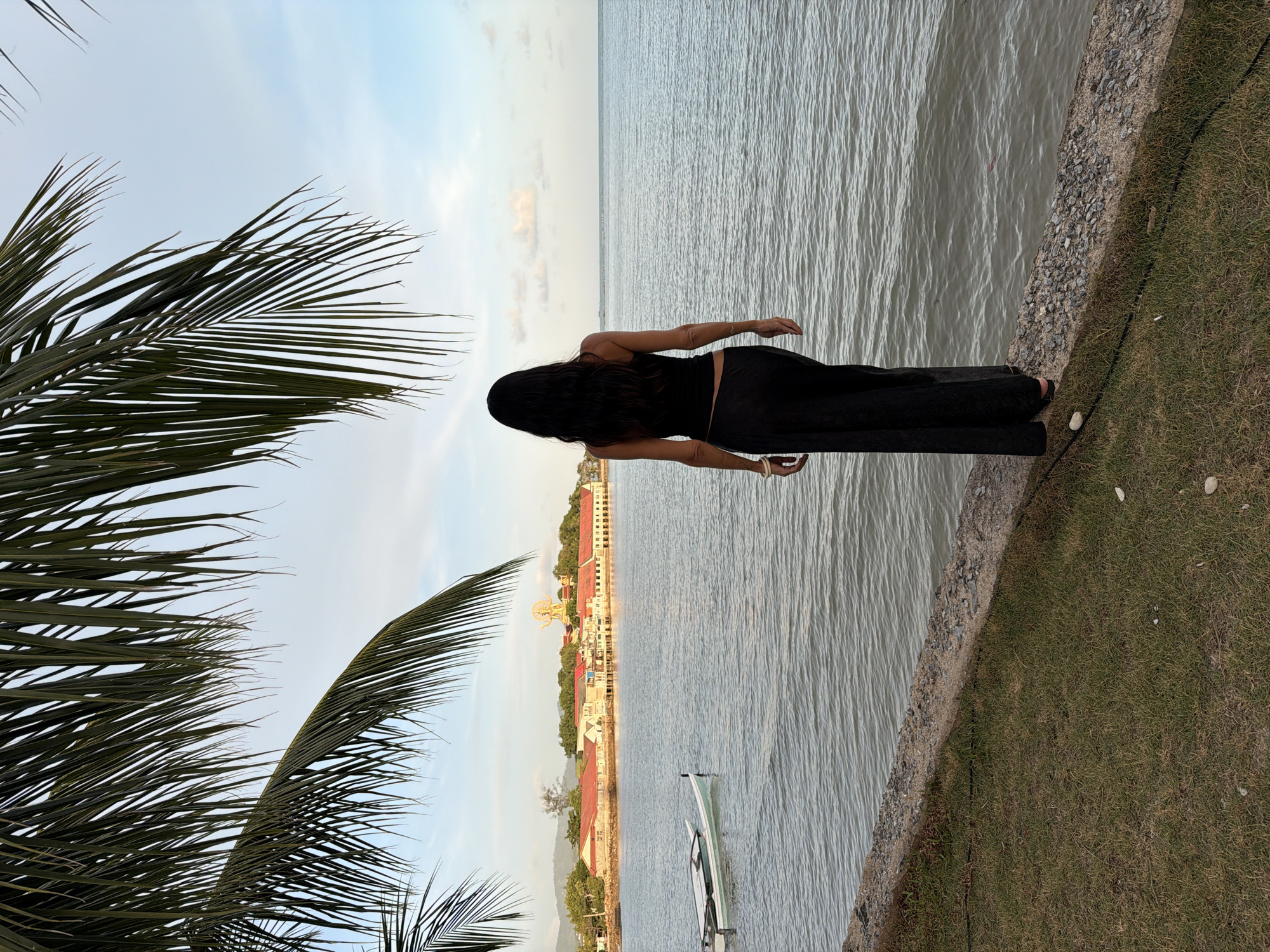 The image depicts a woman standing on a concrete embankment overlooking a body of water. She is viewed from behind, with her dark hair cascading down her back. She is wearing a black tank top and wide-legged black pants. The water is calm with visible ripples. In the distance, across the water, a town is visible, characterized by buildings with red roofs. A prominent golden statue of a deity stands atop a hill behind the town. Two small boats are moored in the water near the shore. Palm fronds hang from the top of the frame, framing the scene. The sky is a light blue with scattered clouds. The area in the foreground consists of grass and the concrete embankment. This scene is consistent with the coastline of the Koh Samui District in Thailand, possibly near a coastal town or village.