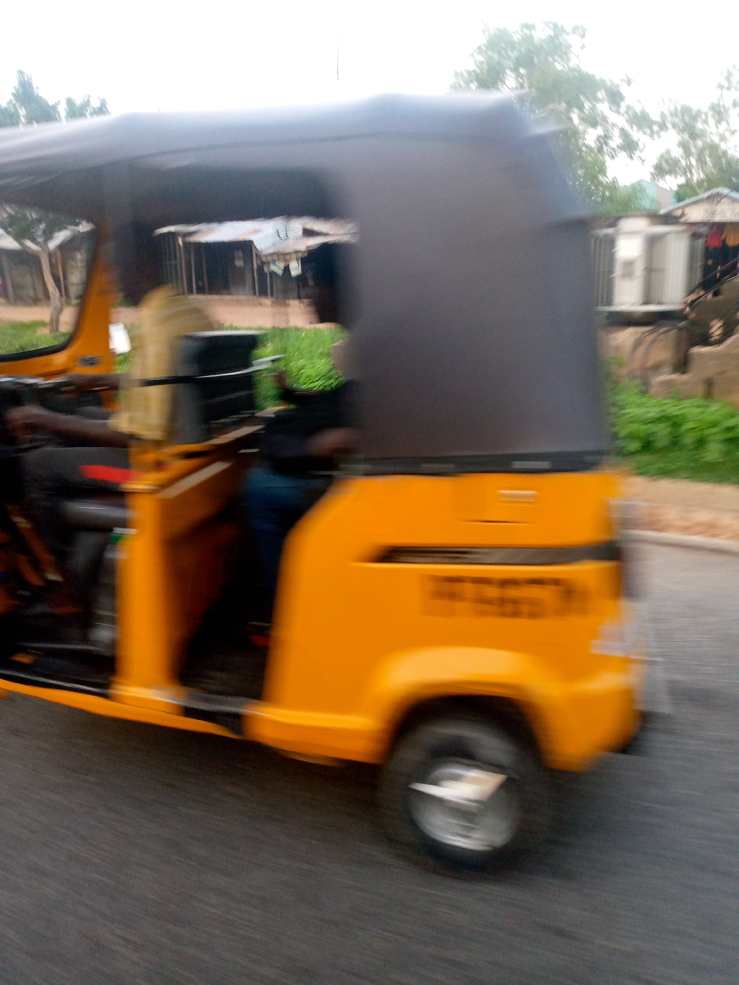 A motion-blurred photograph captures a bright yellow auto-rickshaw, also known as a Keke Napep, traveling on a paved road in Jalingo, Nigeria. The rickshaw has a dark gray fabric roof and a black passenger seat. A person wearing a light-colored shirt is visible in the driver's seat, steering the vehicle. Another person, wearing dark clothing, is seated behind the driver. The background shows a blurred depiction of buildings with corrugated metal roofs and green vegetation. The overall impression is one of movement and a typical urban scene in Nigeria.