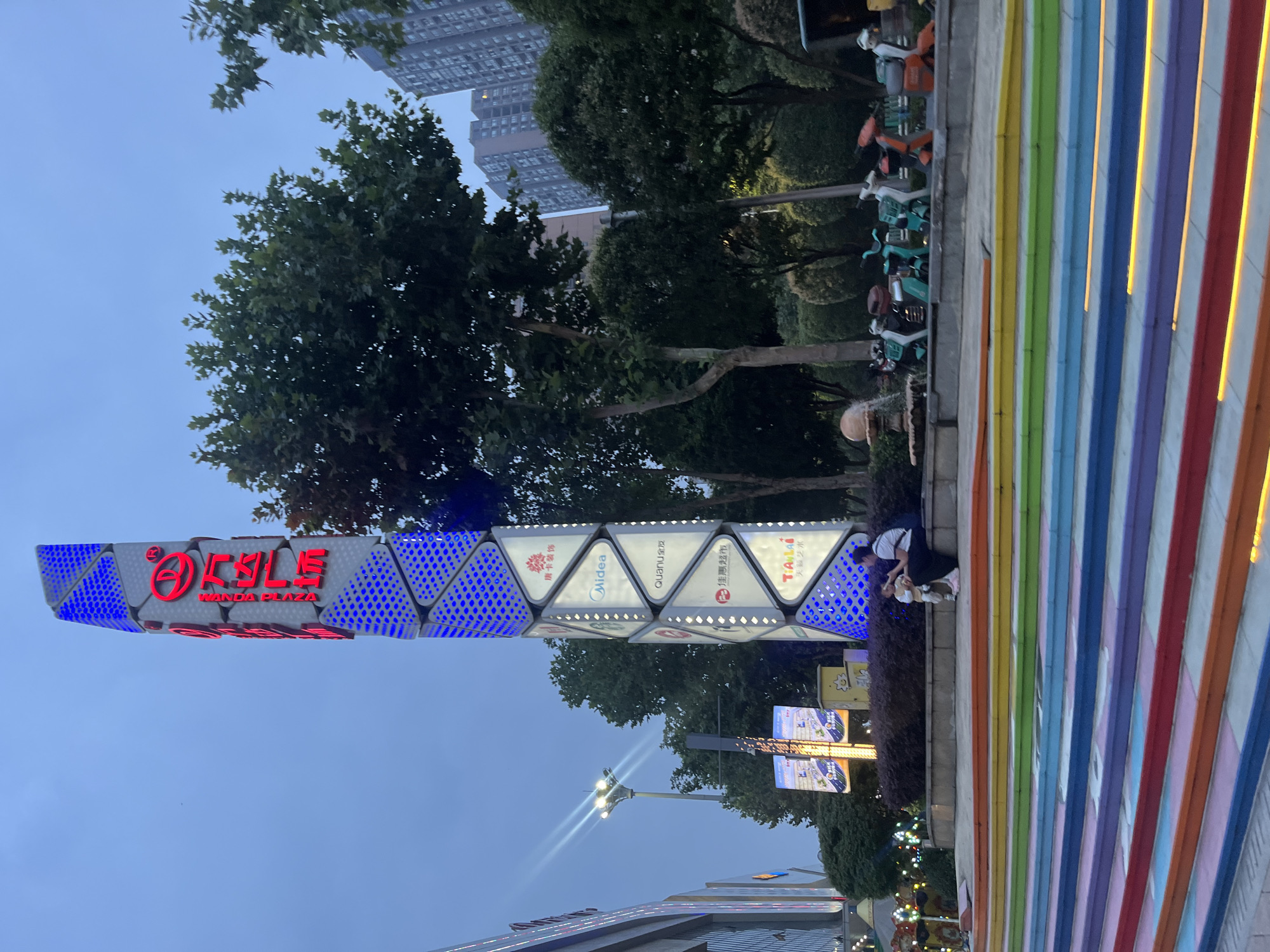 The image depicts an outdoor scene in Shapingba District, China, with a towering vertical sign as the primary focal point. The sign is segmented, with the upper portion displaying red Chinese characters and a circular logo, illuminated by red LEDs. Below this, the sign is adorned with illuminated panels featuring various logos and text, including "Midea," "QuanU," and "Tianlai." The lower part of the sign is patterned with blue LED lights.
In the foreground, colorful, rainbow-striped stairs lead up to a paved area. A woman and a child are seated on a low wall bordering the stairs, interacting with each other. Behind them, a row of small, brightly colored electric scooters is parked. Beyond the scooters, a small fountain is visible, and further in the background, lush green trees and tall apartment buildings rise against a twilight sky. Streetlights are also present, casting a soft glow on the scene.
