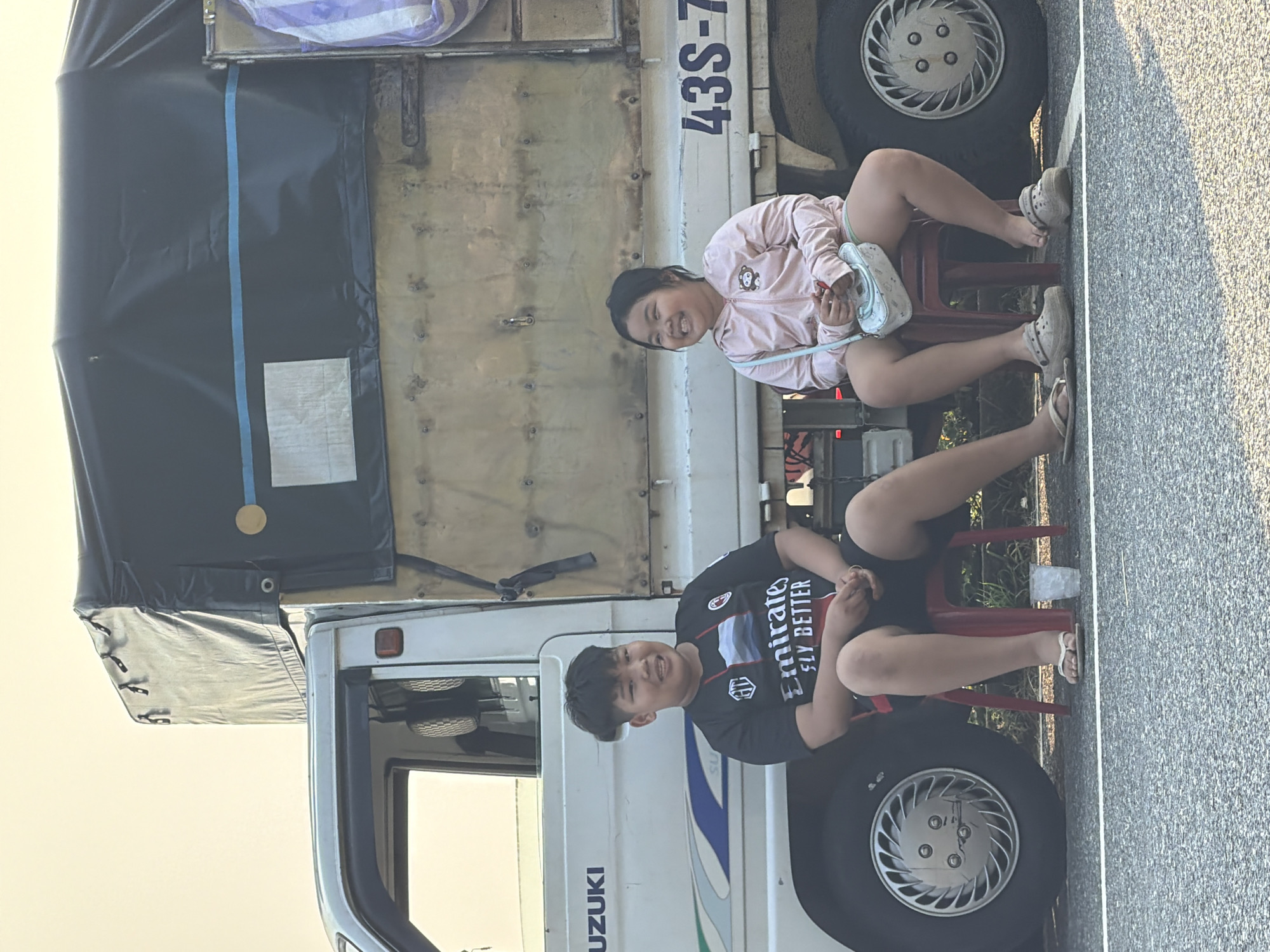 Two children, a boy and a girl, sit on small red plastic chairs next to a Suzuki truck, registration number 43S-7, on a paved road. The girl wears a pink jacket and carries a light-colored purse. The boy wears a black t-shirt with white lettering. They are both smiling. The truck has a dark-colored covered bed with a partially visible white and purple bundle on top. The setting appears to be outdoors, possibly on the outskirts of Đà Nẵng, Vietnam, based on the vehicle registration number.