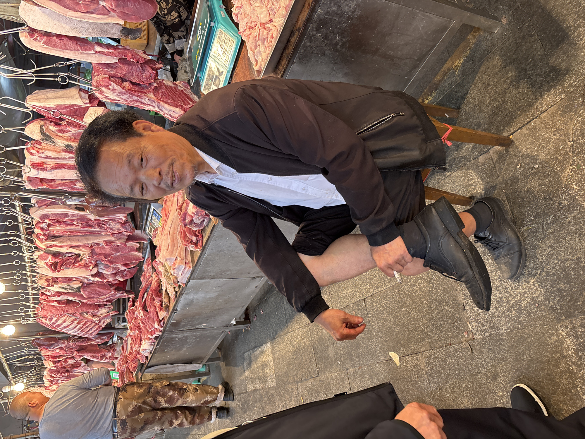 An elderly Asian man sits on a low stool in a butcher shop in Jiangjin, China. He wears a black jacket over a white shirt and black pants. He crosses his right leg over his left and appears to be smoking a cigarette. Fresh meat hangs from hooks behind him. A scale and more meat are visible on the counter next to him. Another man in a gray shirt and camouflage pants is partially visible in the lower left of the image. Two people wearing black are present in the lower part of the image. The floor is concrete.