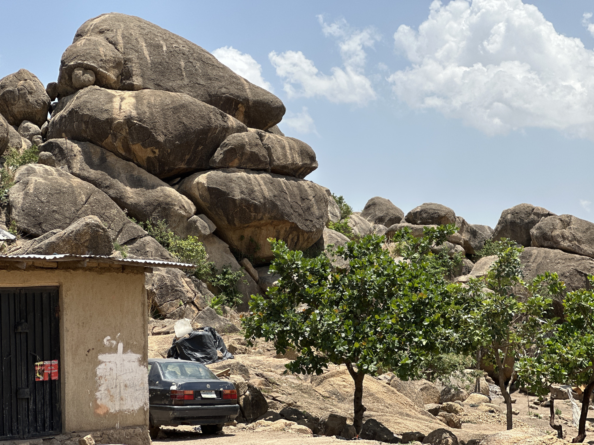 The image shows a rocky landscape with a building and a car, likely located in Jos, Nigeria, given the terrain. The foreground features a small, rough-hewn building with a corrugated metal roof and a black door with a sign. Beside the building is a dark-colored car, parked on a dirt surface. Adjacent to the car, a black plastic bag is visible. The background consists of large, rounded rock formations typical of the Jos Plateau, interspersed with green vegetation. The sky is partially clear with some scattered clouds. The overall scene appears to be a rural or semi-rural area with signs of human habitation and transport.