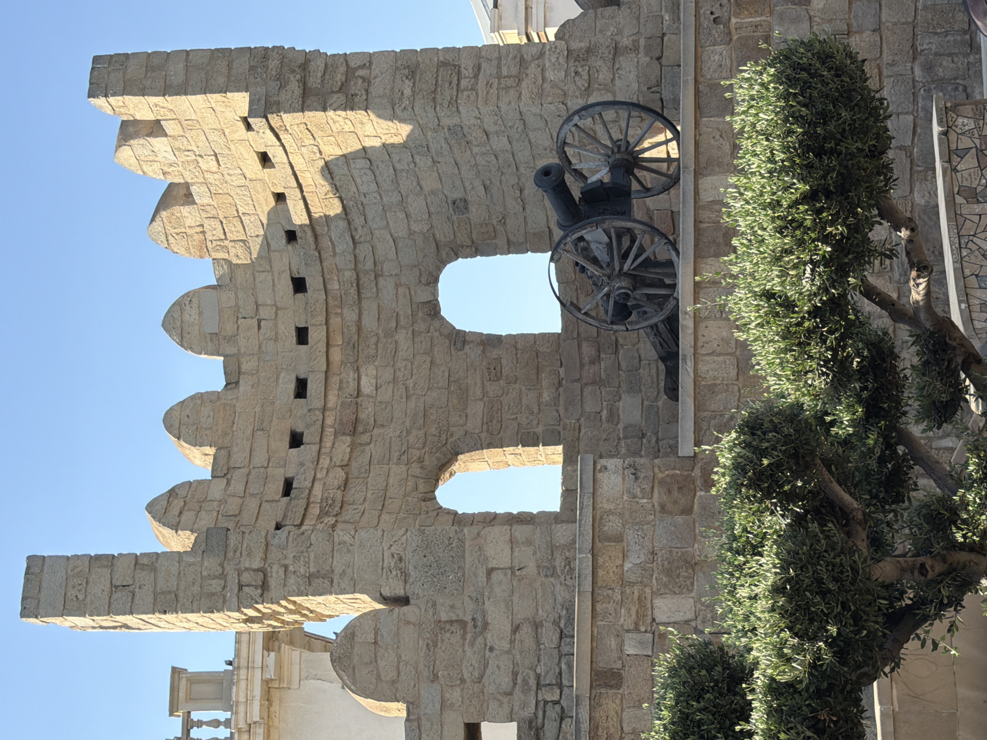 This image depicts a historical stone structure, likely a part of the Old City of Baku in Sabail Raion, Azerbaijan. The structure is made of light-colored stone blocks and features crenellations at the top, resembling battlements. There are two arched openings visible within the wall. In front of the wall, a dark-colored cannon with wooden wheels is positioned. In the foreground, several well-maintained green bushes and a tree with visible branches are present, partially obscuring the lower portion of the stone wall. The sky is clear and blue. The scene appears to be outdoors during daylight hours.