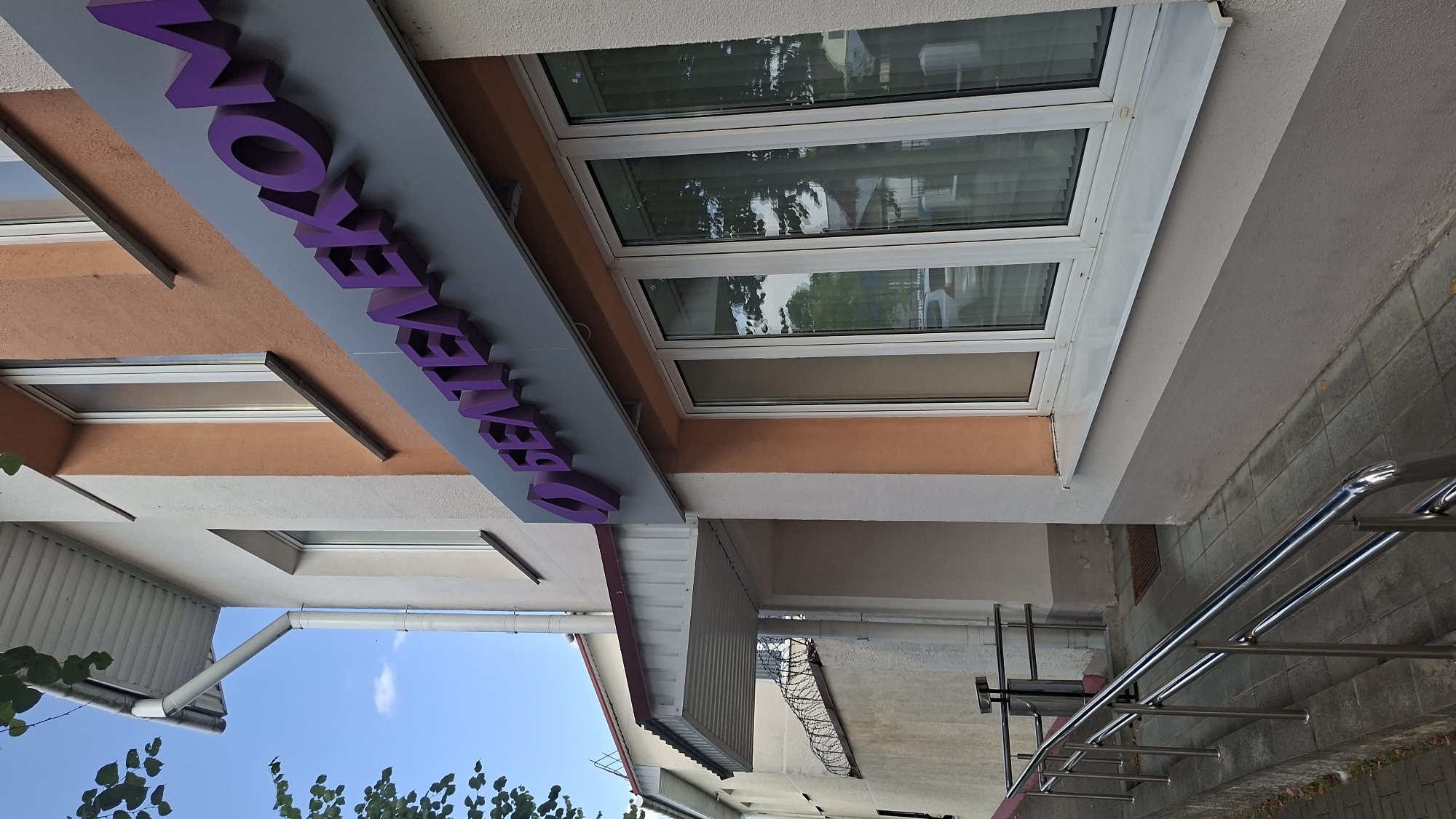 The image shows the exterior of a building in Myadzyel, Belarus, with a sign reading "БЕЛТЕЛЕКОМ" in purple lettering. The building has a light beige facade and an orange accent wall. There are several windows on the building, some of which reflect trees and vehicles. A metal ramp with handrails leads up to the entrance. A barbed wire fence is visible on the side of the building, suggesting a secure area. The sky is visible in the upper left corner, with some green foliage in the foreground. The overall scene depicts a commercial establishment, likely a telecommunications office, in a suburban or semi-urban setting.